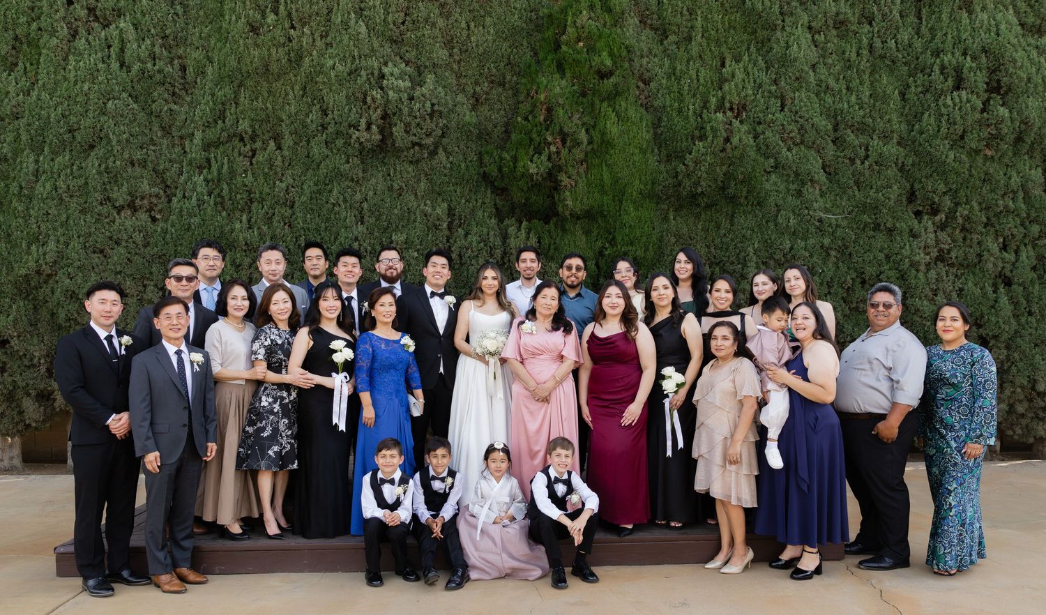 Large family group posed outdoors for a formal wedding celebration photo in front of a lush green hedge wall.