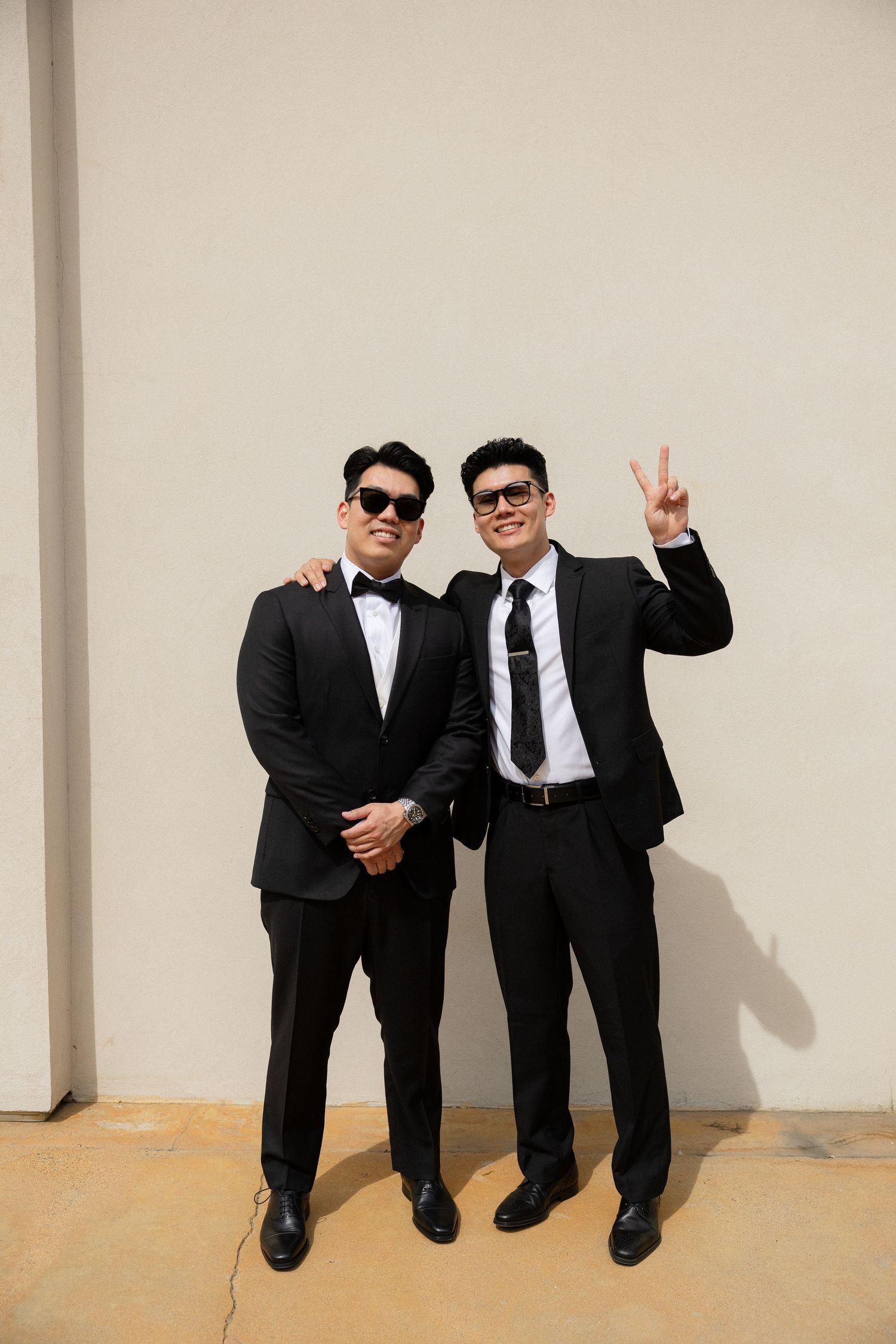 Two stylish men in black suits and sunglasses posing outdoors, one making a peace sign.