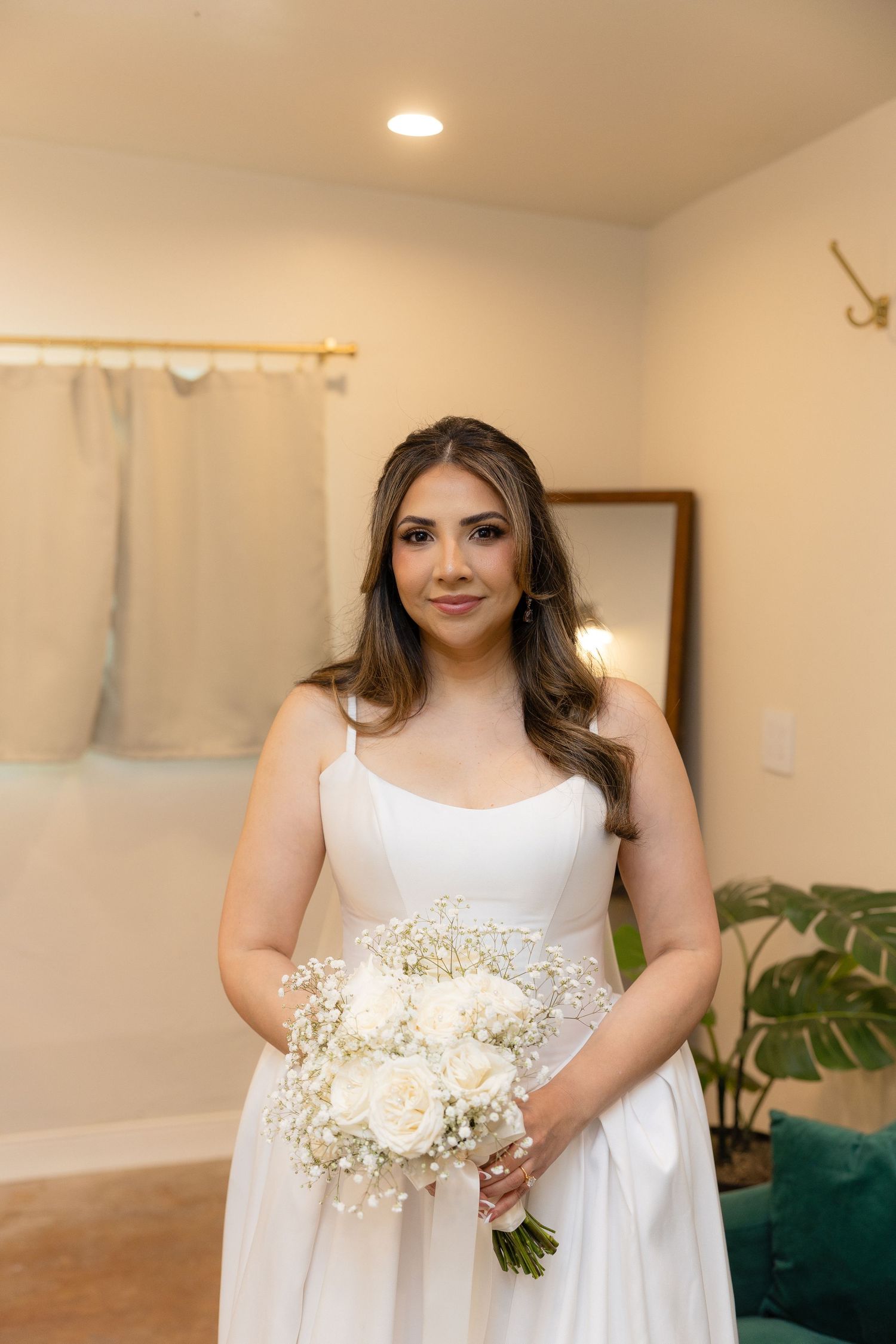 Bride in white gown holding white rose and baby's breath bouquet poses indoors before wedding ceremony.