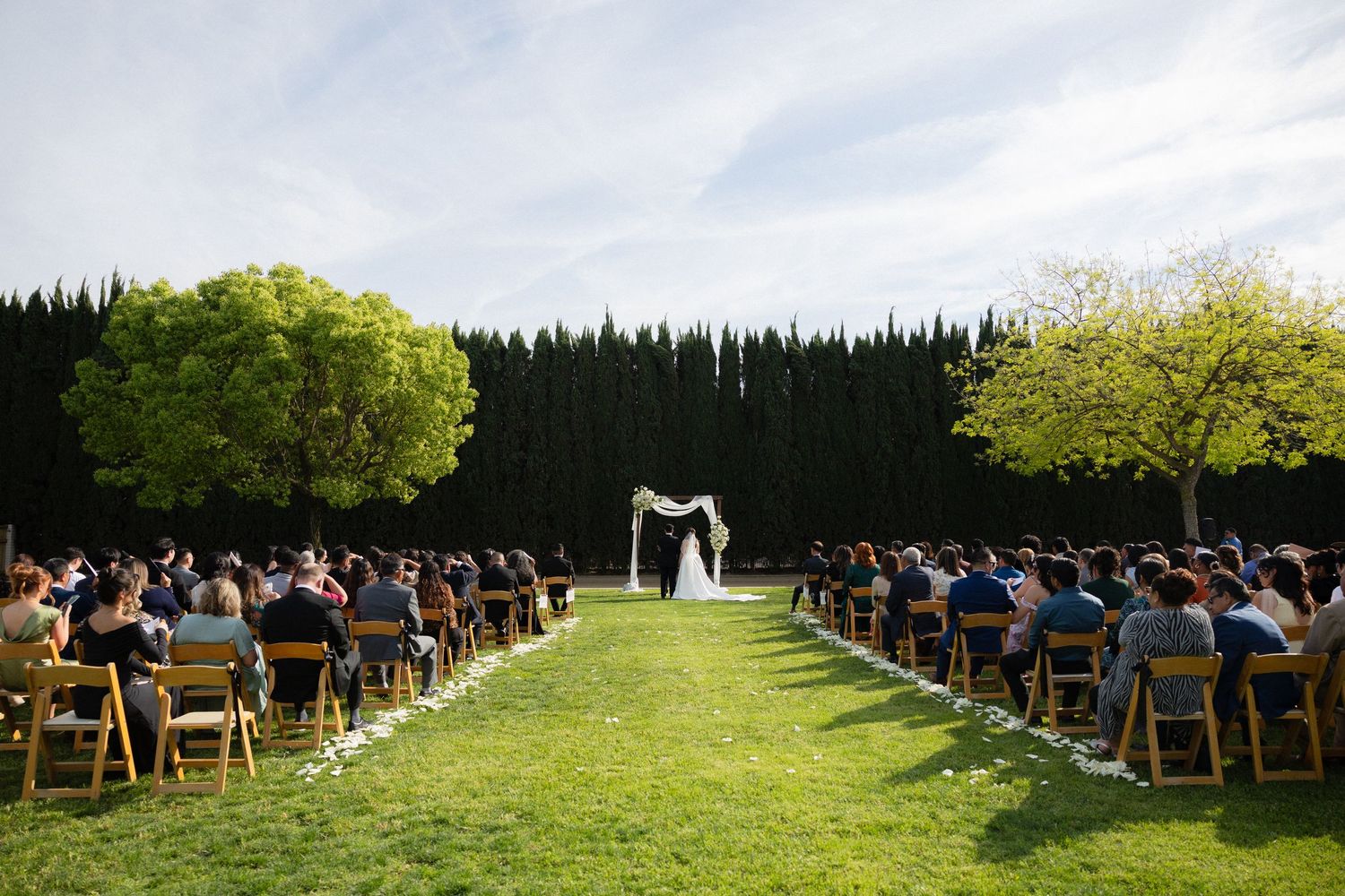 Outdoor wedding ceremony with guests seated in wooden chairs facing a white floral arch.