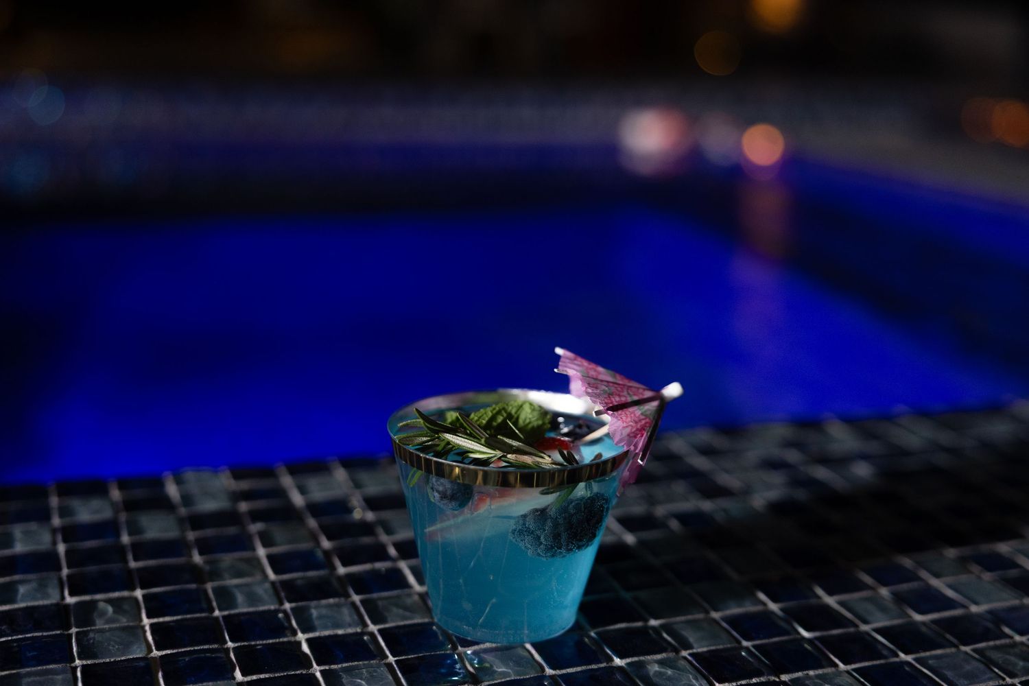 A blue cocktail garnished with fresh herbs sits on a dark tiled surface with vibrant blue lighting in the background.
