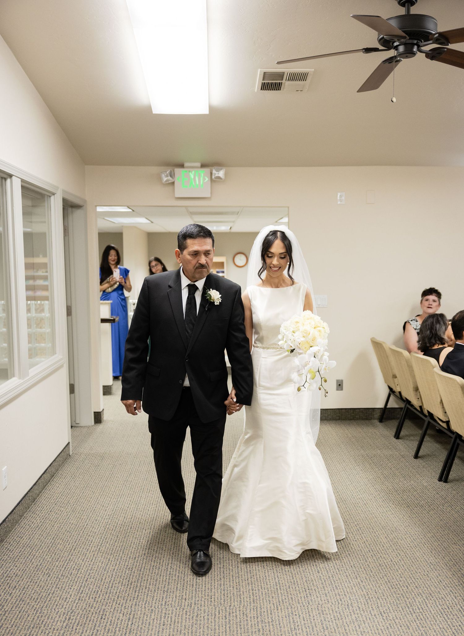 A wedding couple and guests walk through a bright hallway with beige carpeting and white walls.