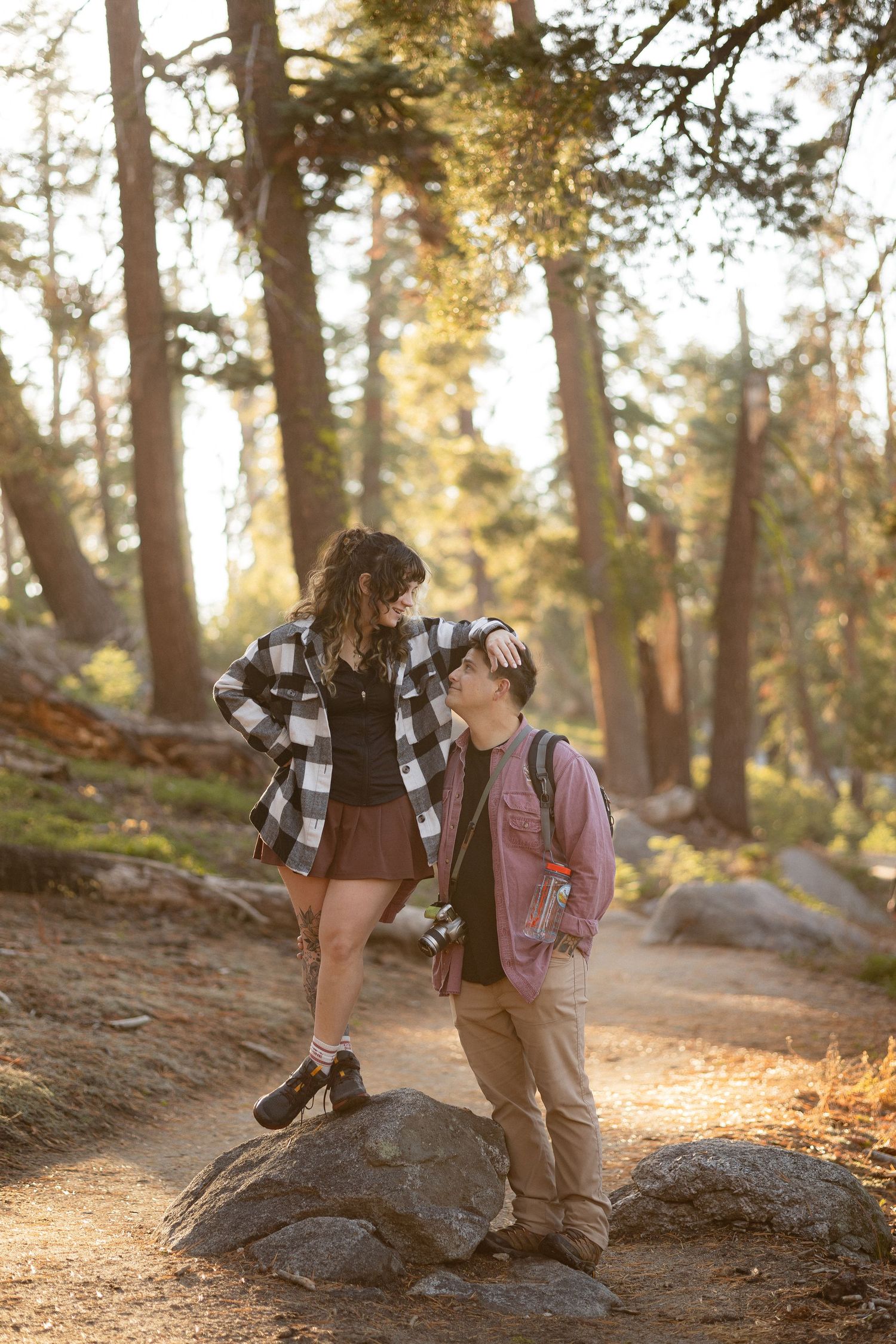 Two people sit together on a large boulder in a sunlit forest setting.