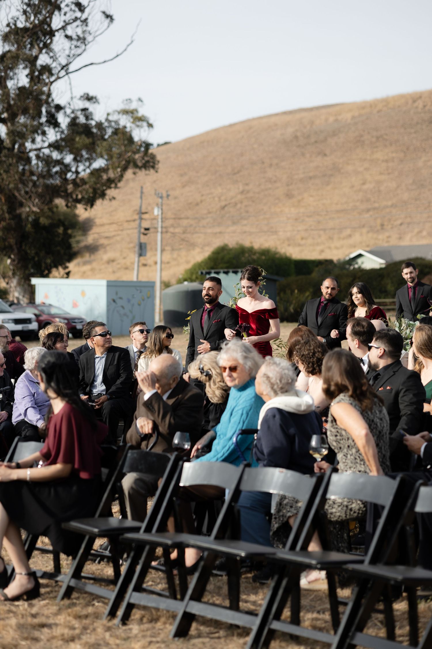 Wide shot of wedding guests gathered for outdoor ceremony in rural setting.