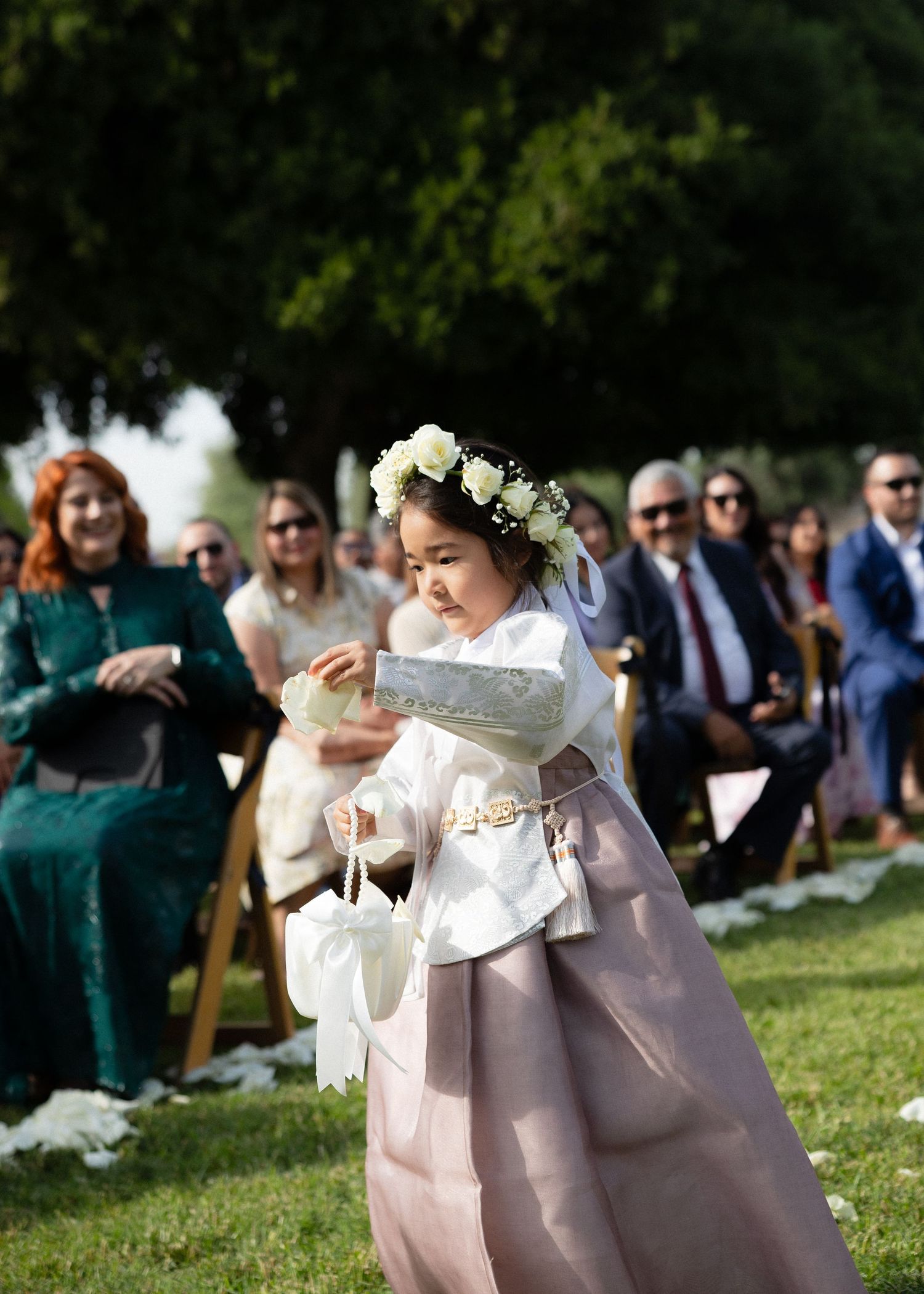 Young flower girl in a traditional Korean hanbok with floral crown scatters petals at an outdoor wedding.