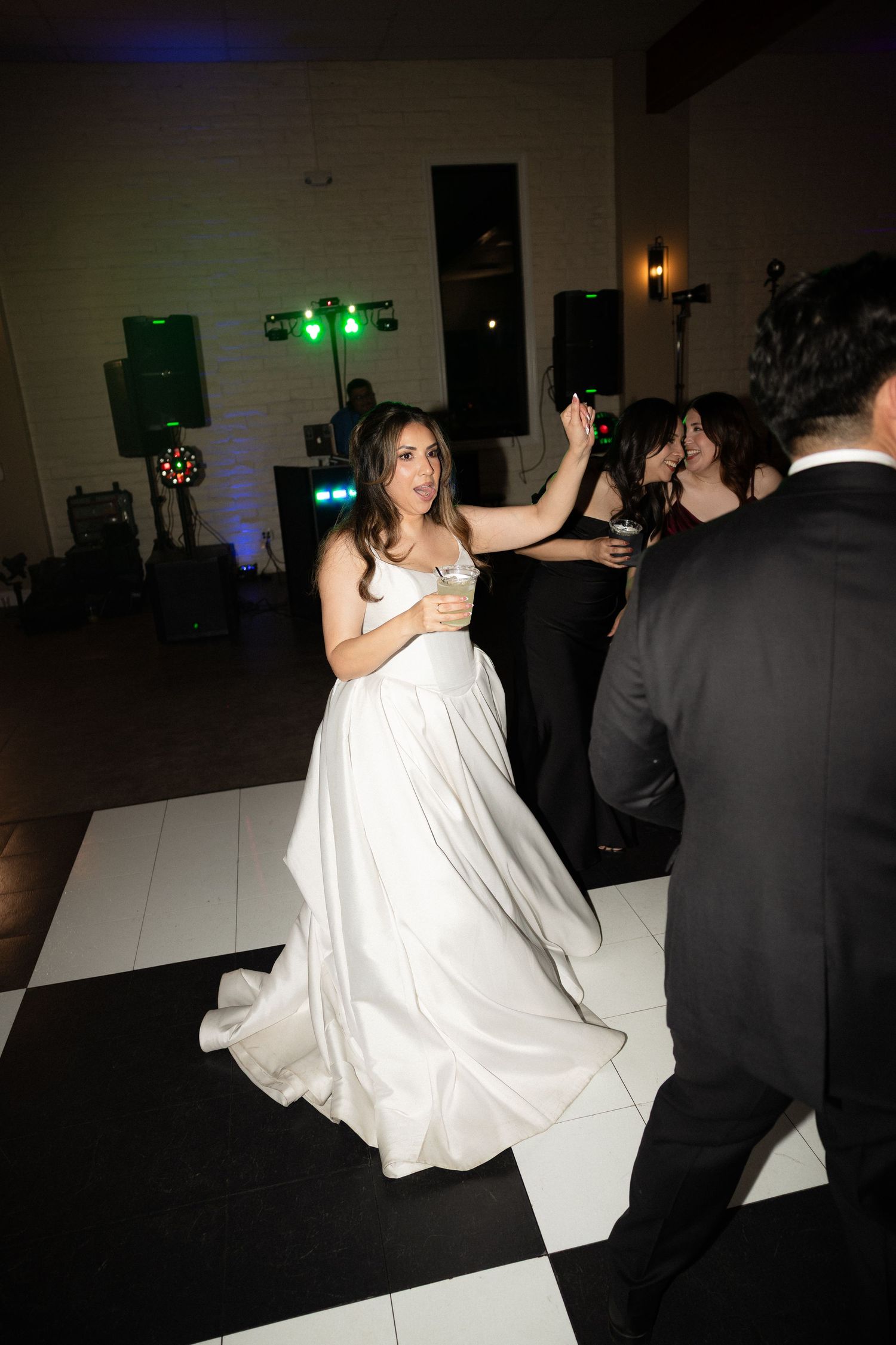 Bride in flowing white gown dances joyfully holding drink on checkered dance floor at reception.