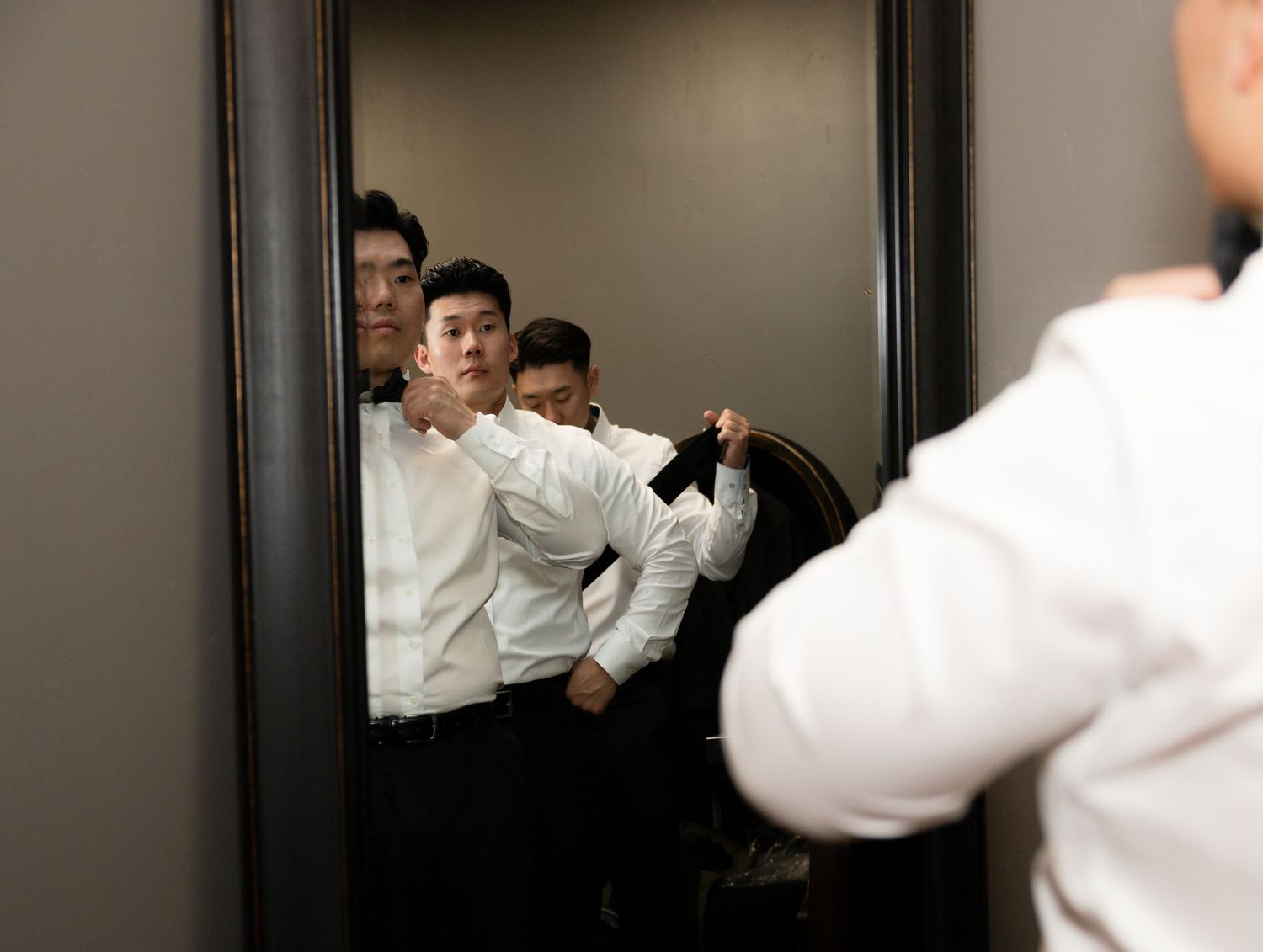 Asian groomsmen in white dress shirts adjusting ties and suit jackets while preparing in front of a large mirror.