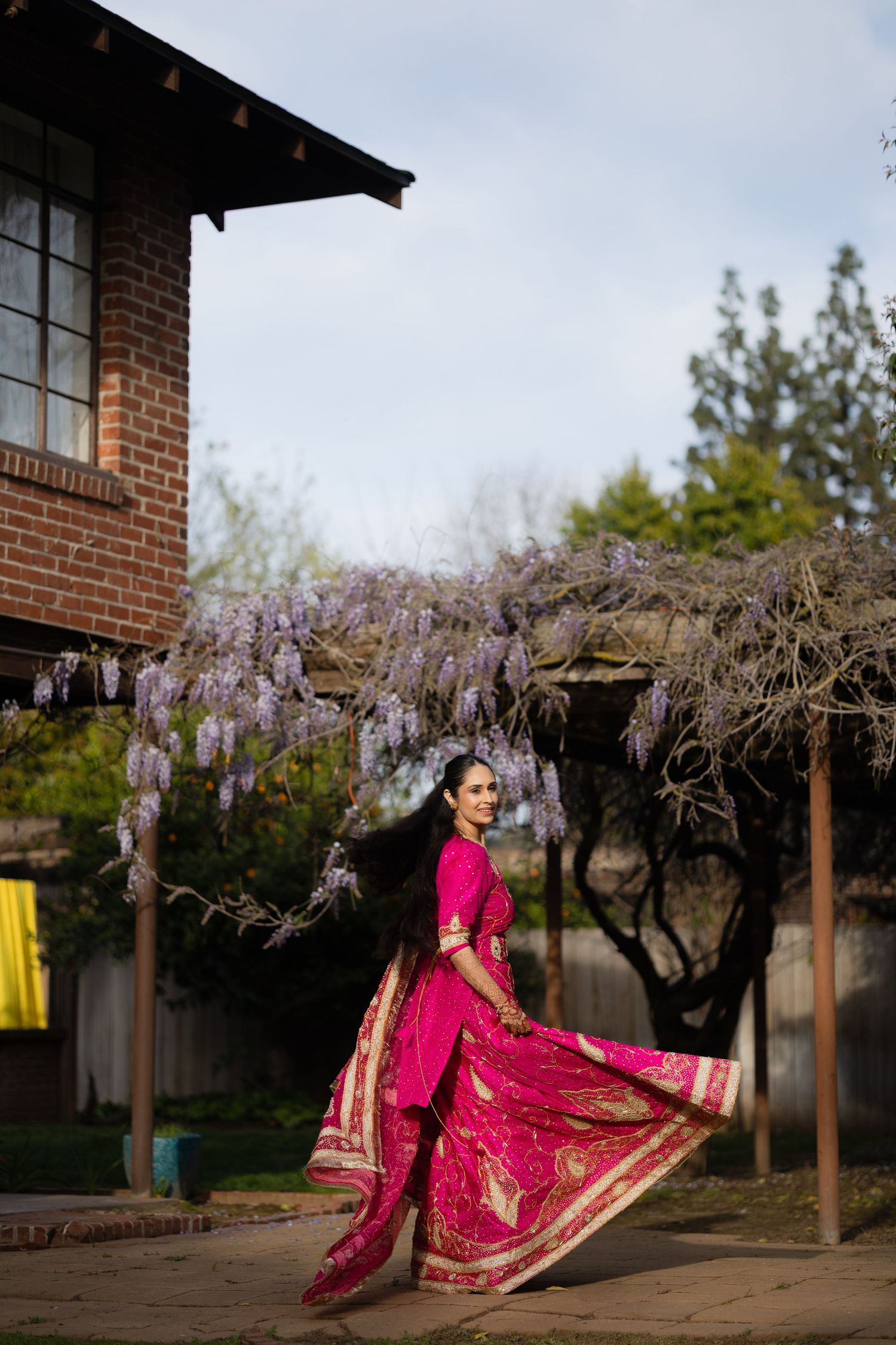 Vibrant pink and gold Indian wedding dress flows gracefully under blooming wisteria trees in an outdoor garden setting.