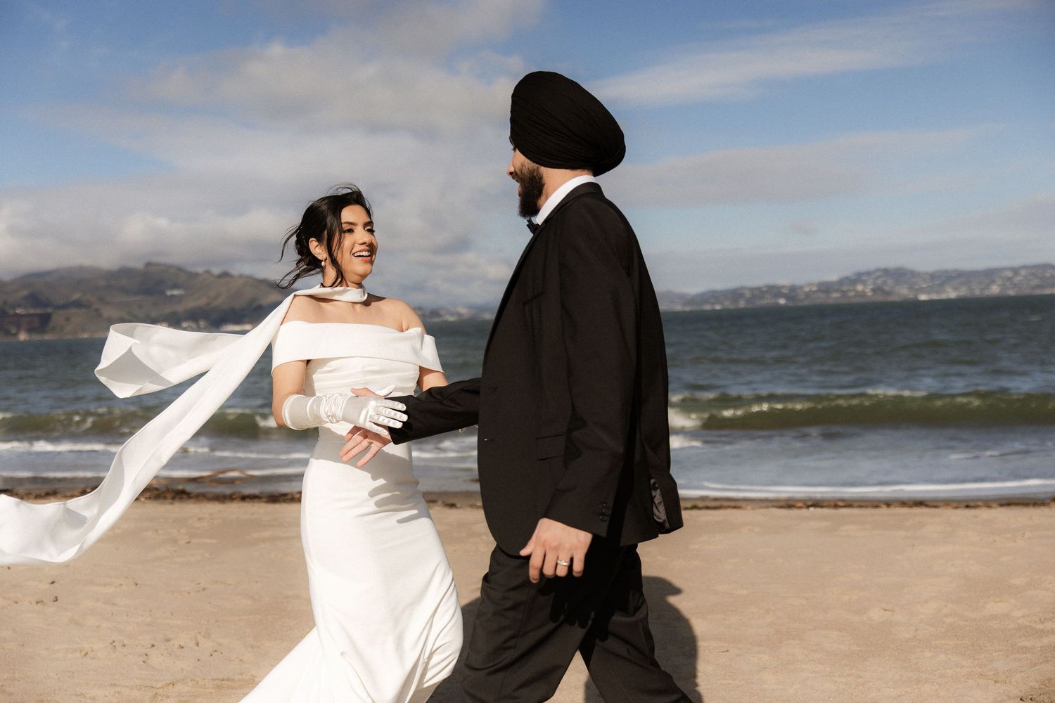 A couple in wedding attire dances together on a beach with mountains and ocean waves in the background.