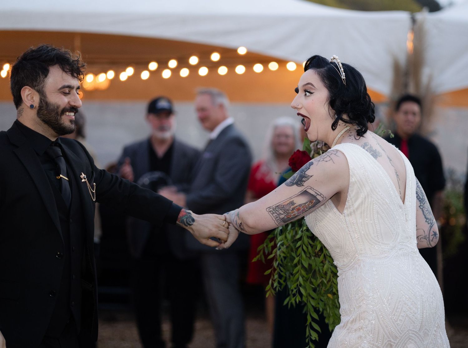 Guests dance and celebrate at an outdoor wedding reception under a tent with string lights.