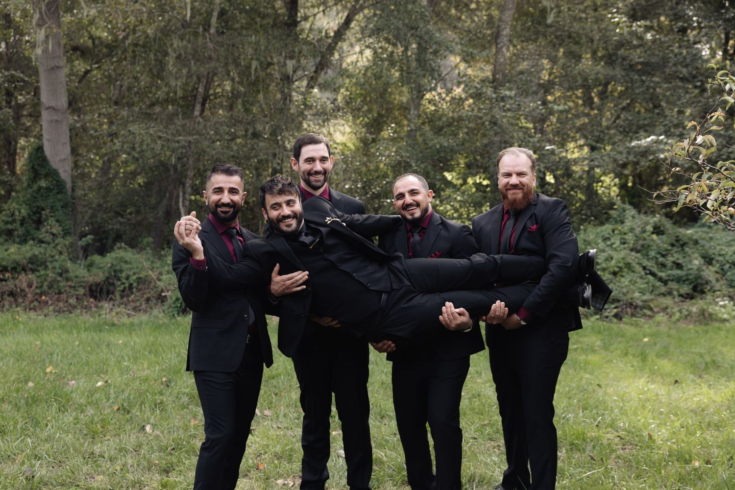 Groomsmen in black suits posing casually outdoors with natural backdrop.