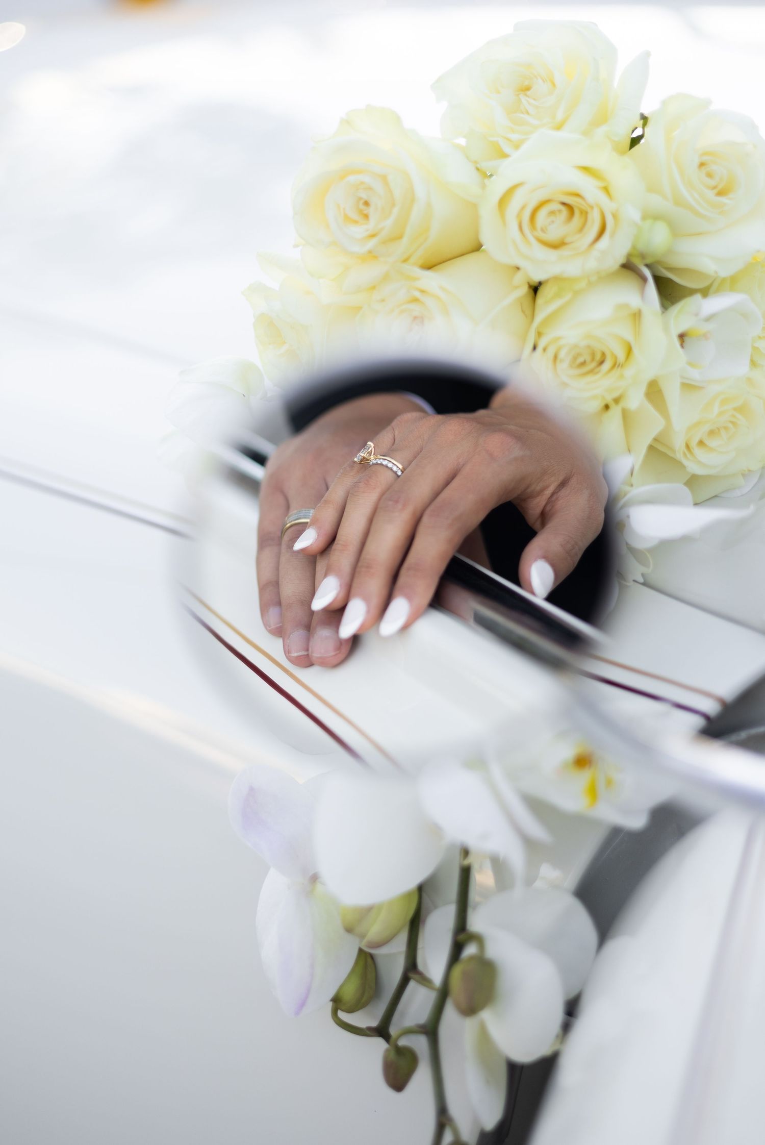 Close-up of hands resting on white vintage car with cream roses in background.