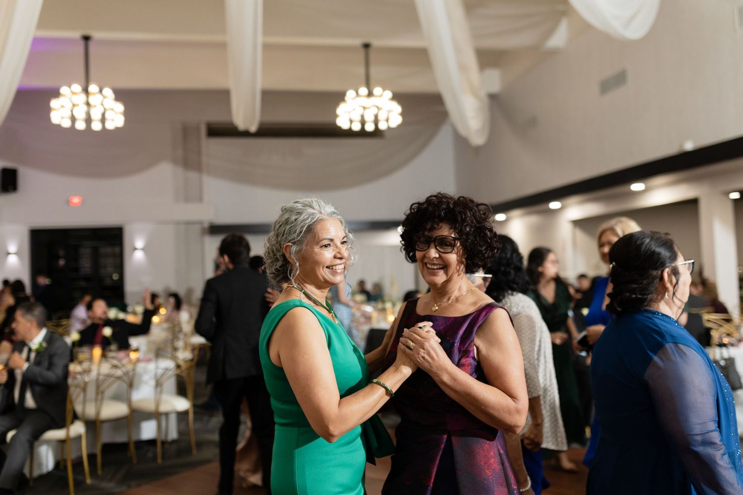 Wedding guests dance and celebrate together under elegant chandeliers at a reception venue.