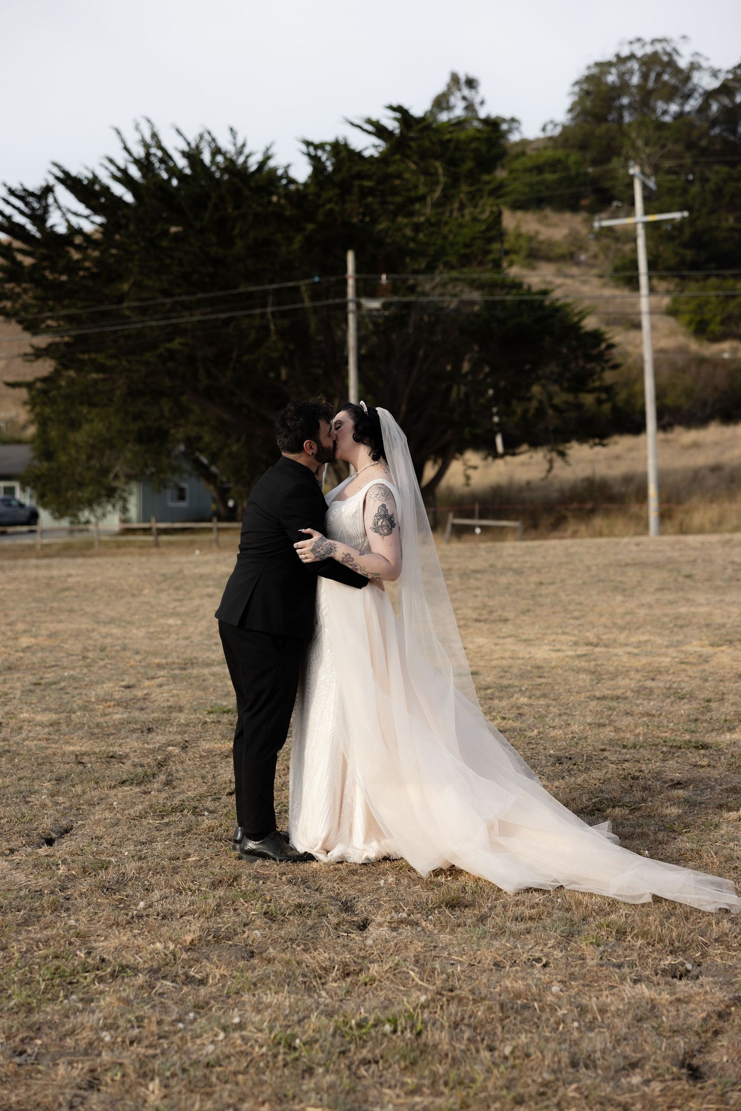 A wedding couple shares an intimate moment in a field with utility poles and cypress trees in the background.