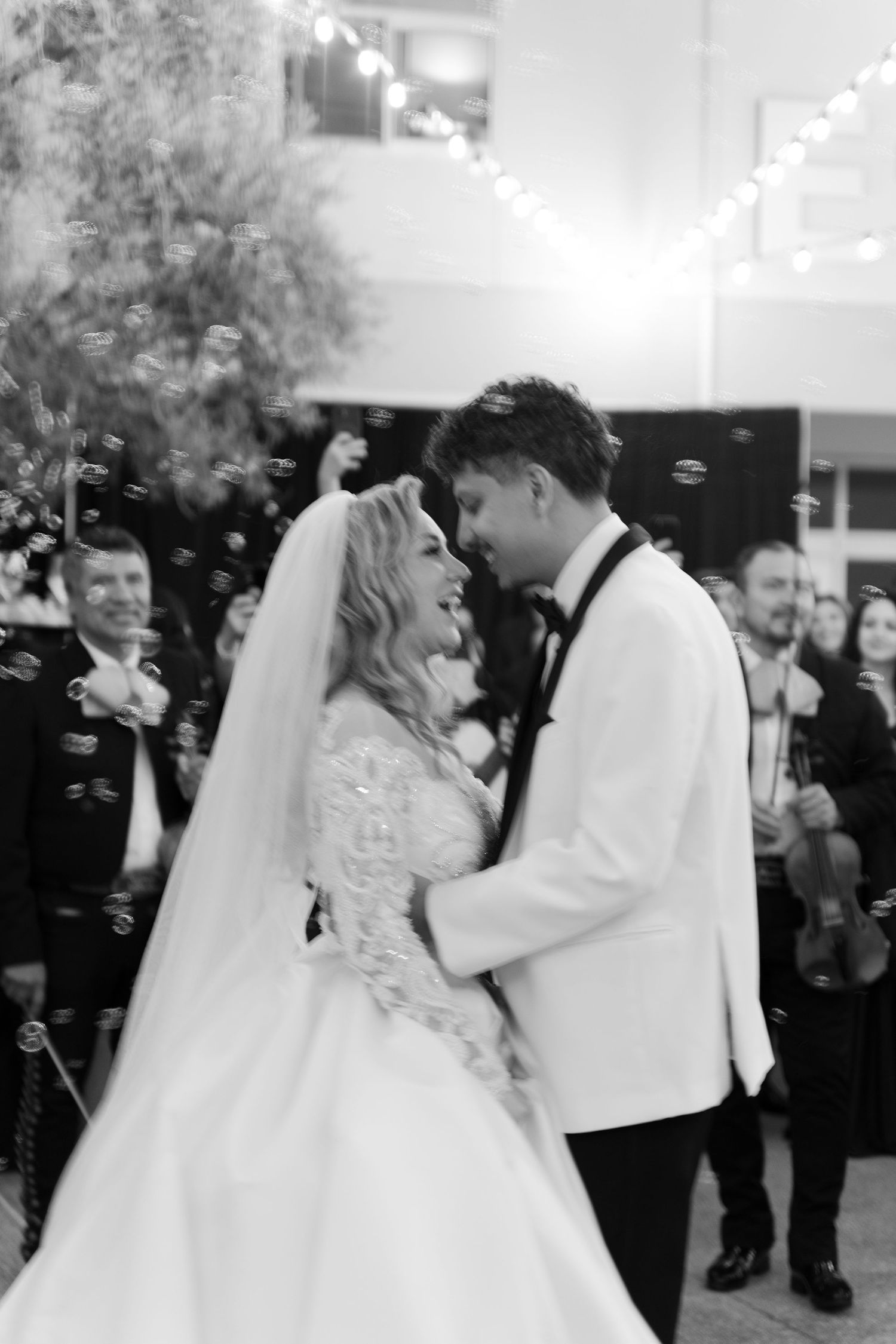 A bride and groom share an intimate dance surrounded by wedding guests and string lights.