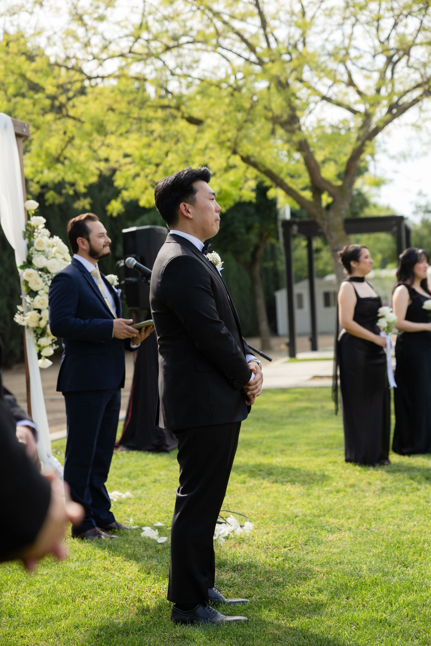 Groom standing at outdoor wedding altar on green lawn with officiant and bridesmaids in black dresses nearby.