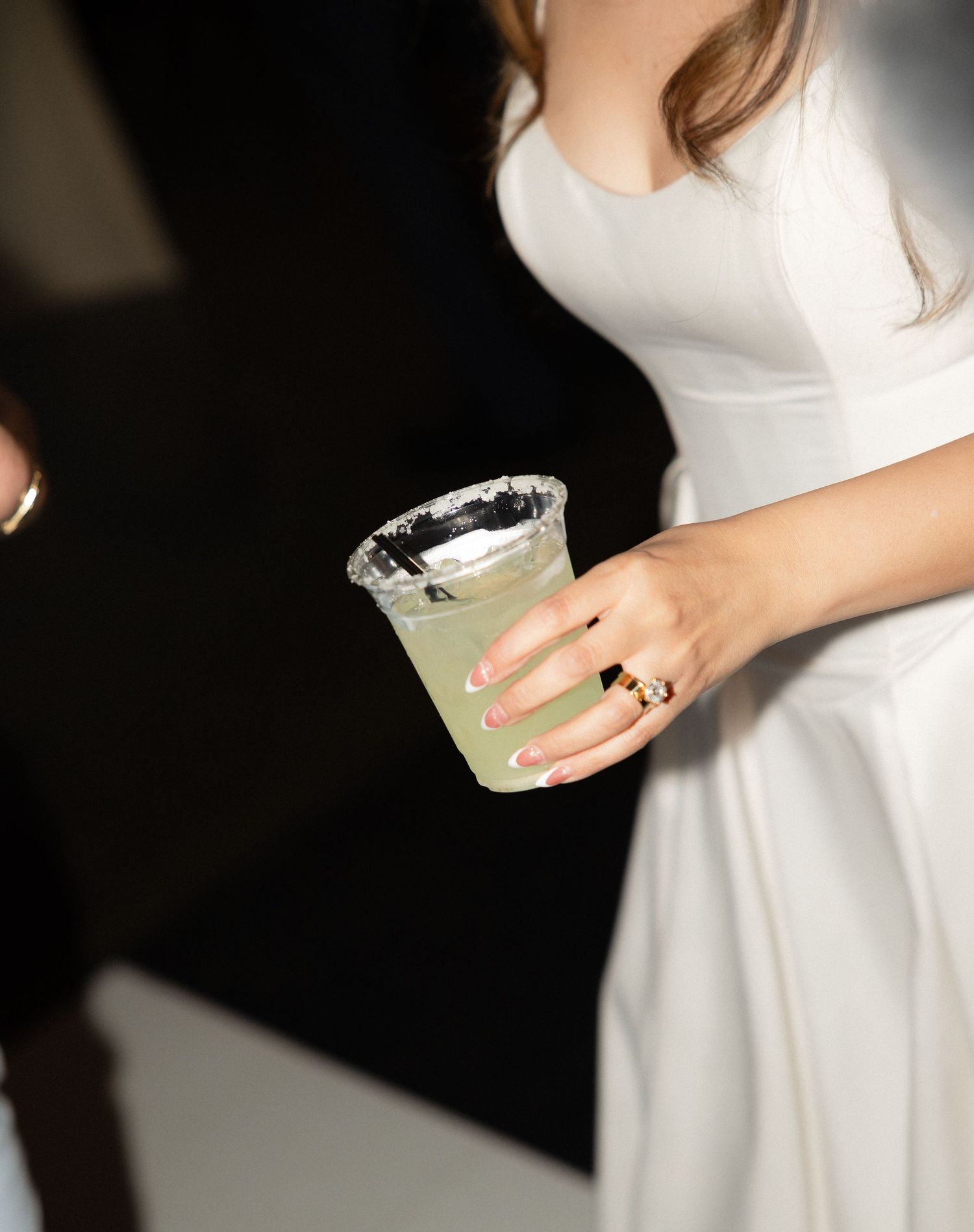Bride in white dress holds salted rim cocktail glass with engagement ring visible.