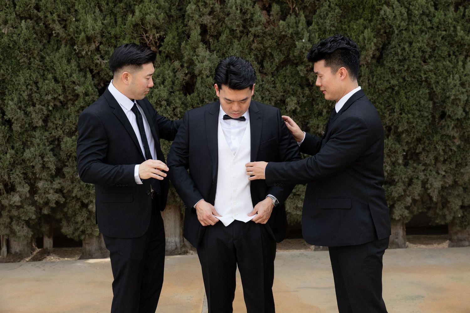Three groomsmen in black tuxedos helping groom adjust his formal attire outdoors before wedding ceremony.