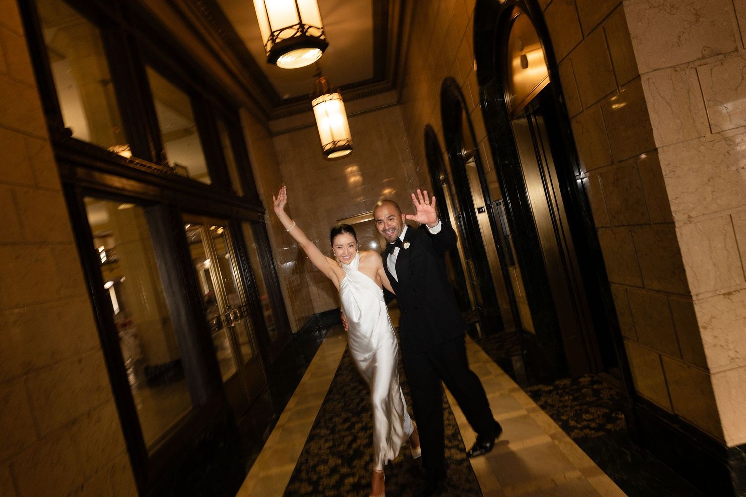 A couple in wedding attire dances joyfully down an elegant hallway with warm lighting and stone columns.