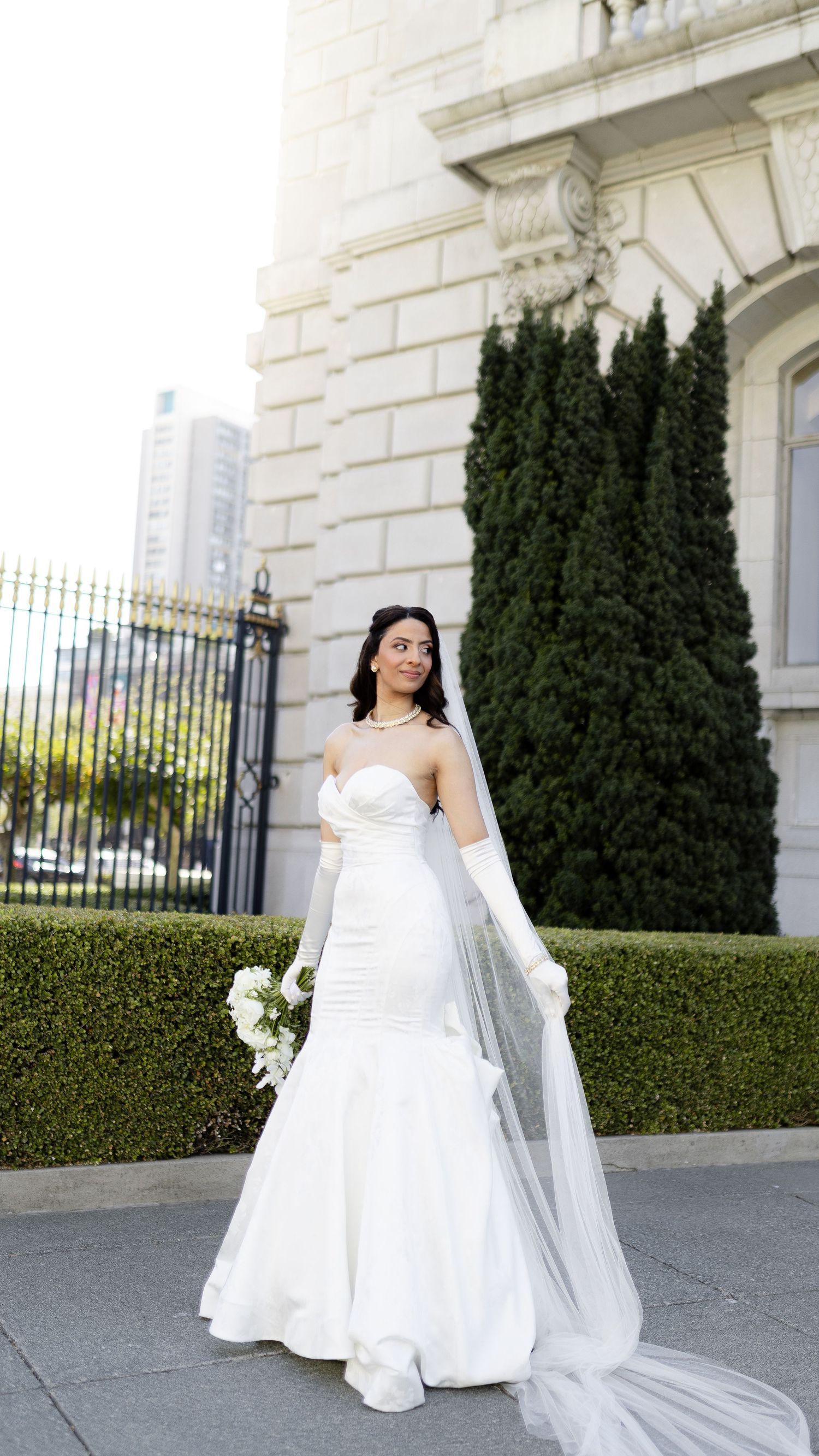 A bride in a white strapless gown poses elegantly against a classic building exterior with manicured hedges.