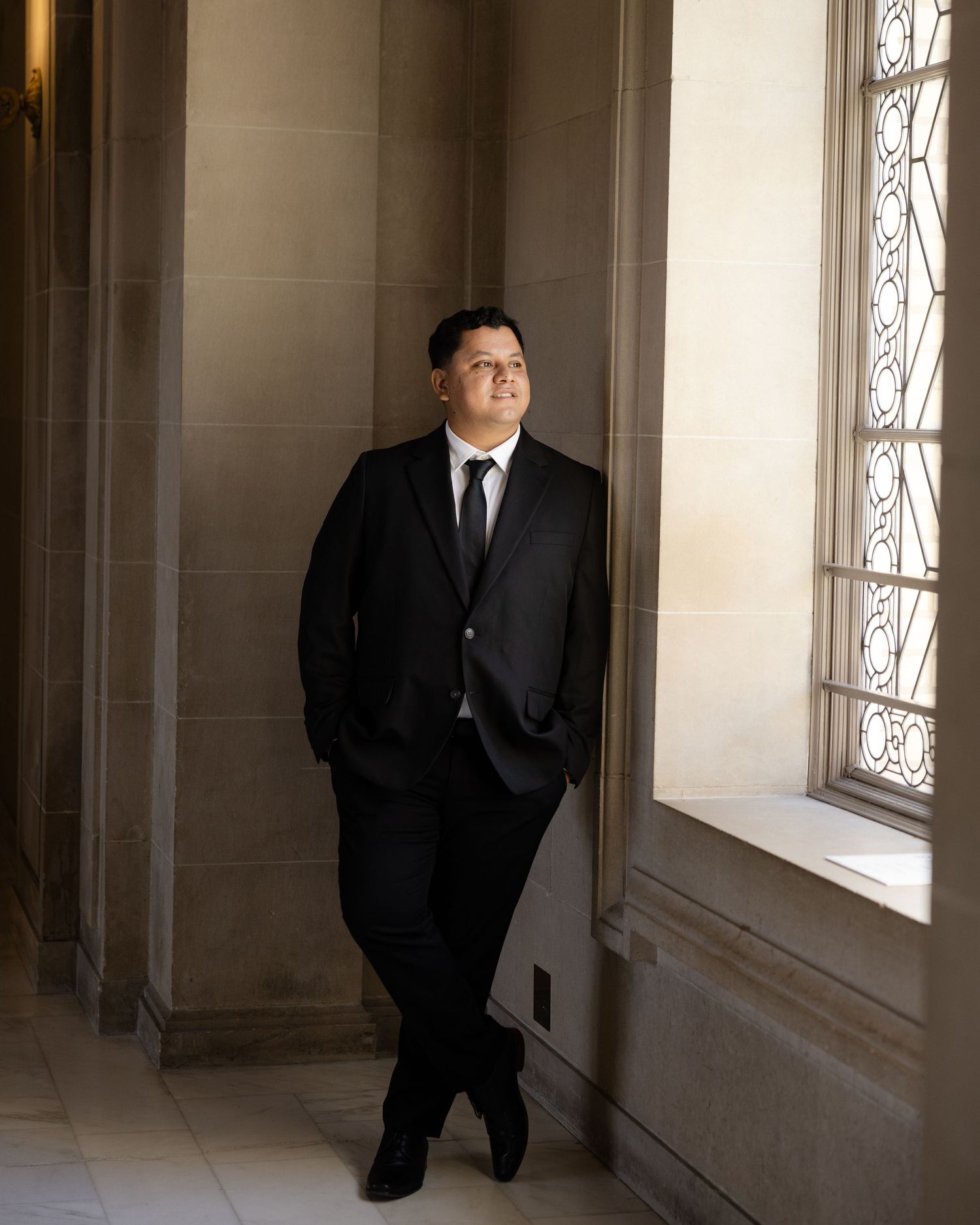 A person in a black suit poses against a bright window in an elegant hallway with white walls.
