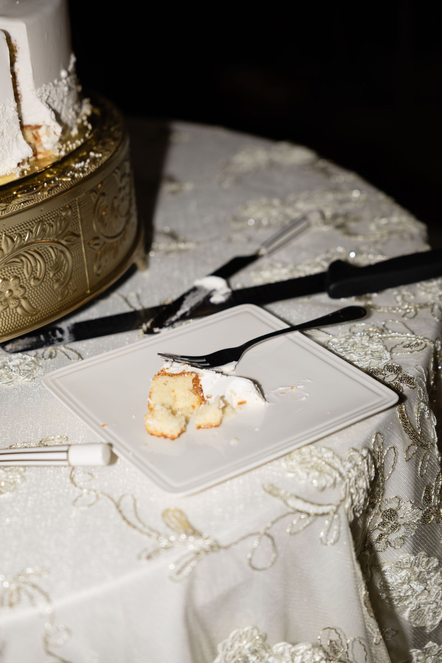 Wedding cake cutting setup with gold cake stand and silverware on white linen tablecloth.