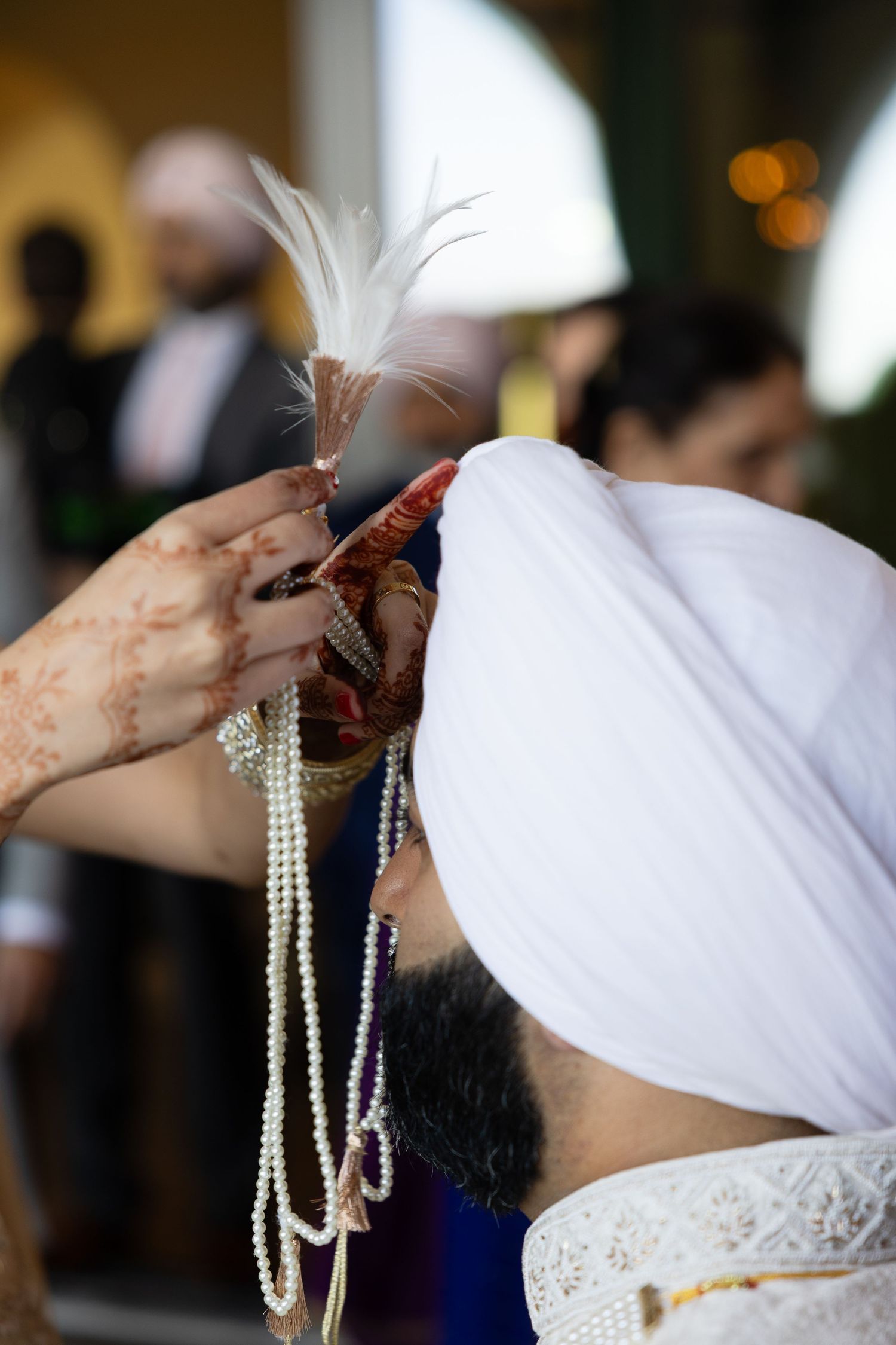 Someone wearing a white turban and traditional dress holds a decorative feather and beaded necklaces during a ceremony.
