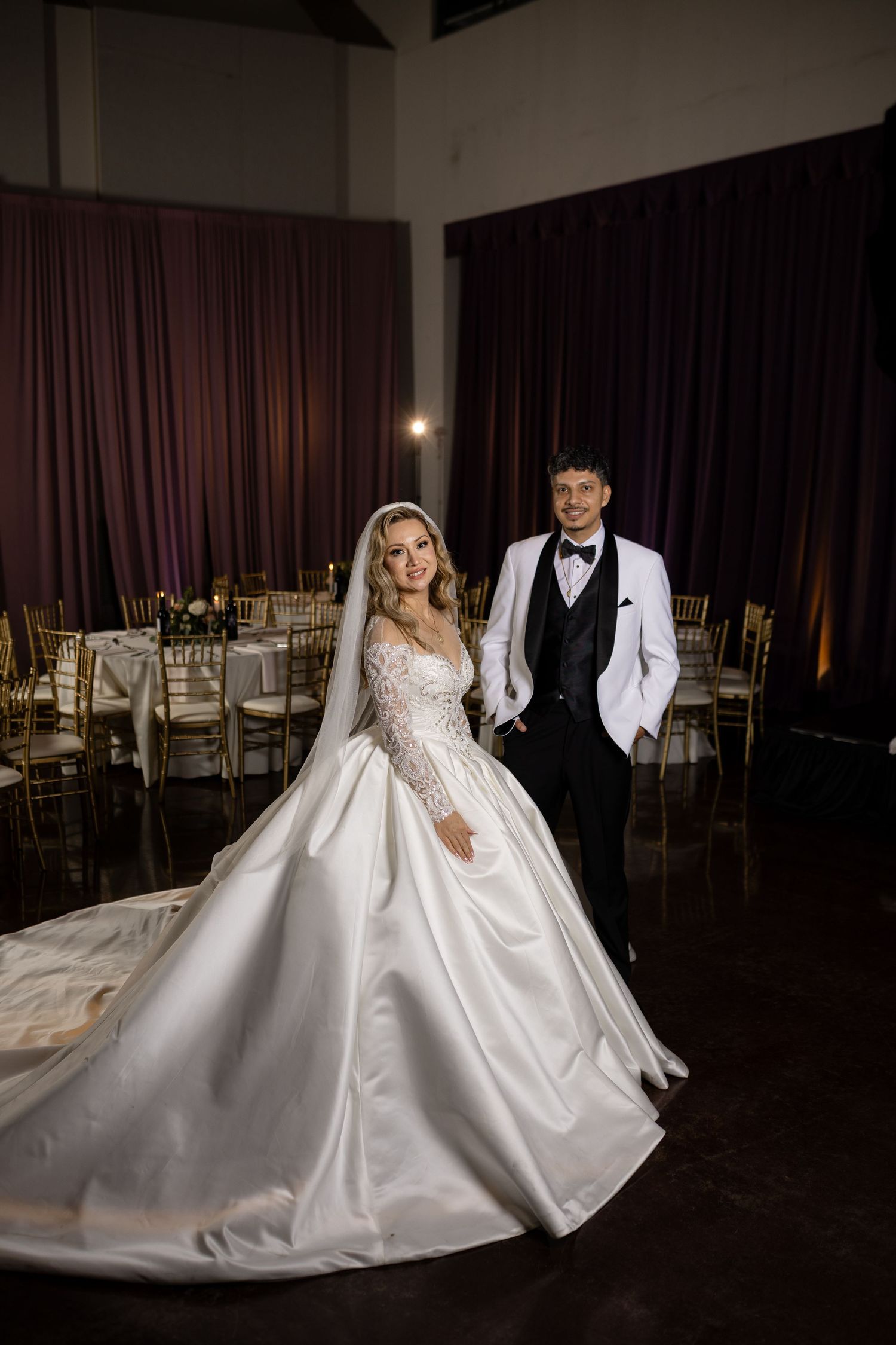 An elegant wedding couple poses dramatically in a ballroom with flowing white dress and tuxedo.