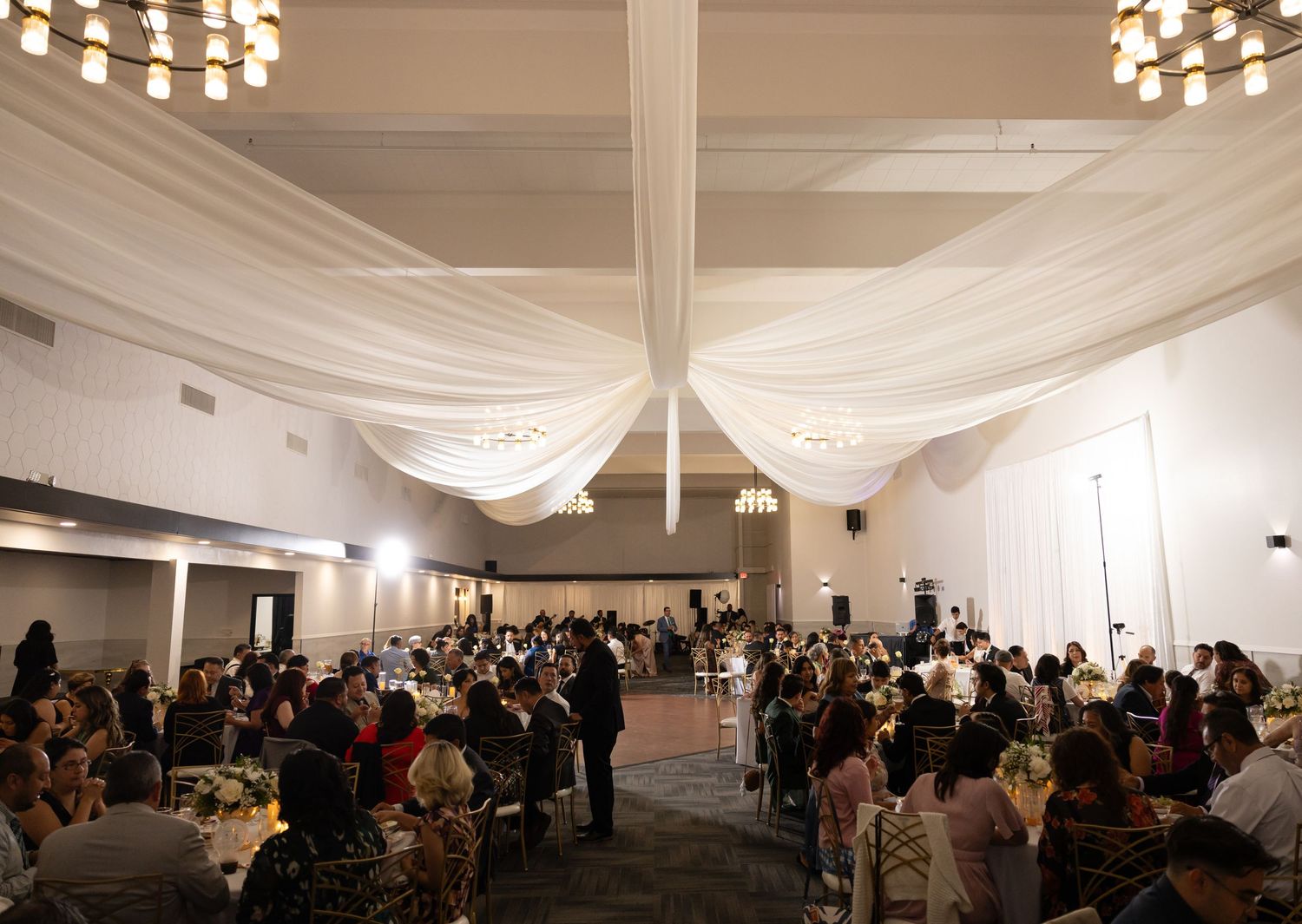 Large wedding reception hall with draped ceiling and chandeliers filled with guests at round tables.
