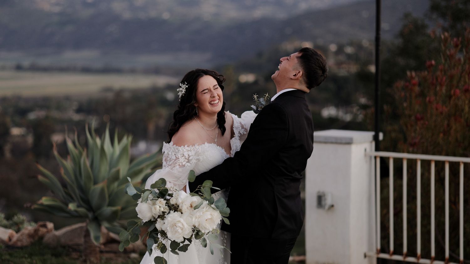 A romantic wedding couple embraces on a balcony overlooking a scenic mountain landscape at sunset.