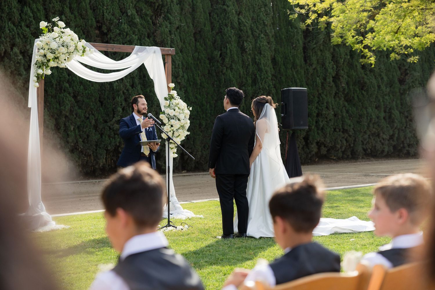 Bride and groom exchange vows at a floral arch during an outdoor wedding ceremony with seated guests.