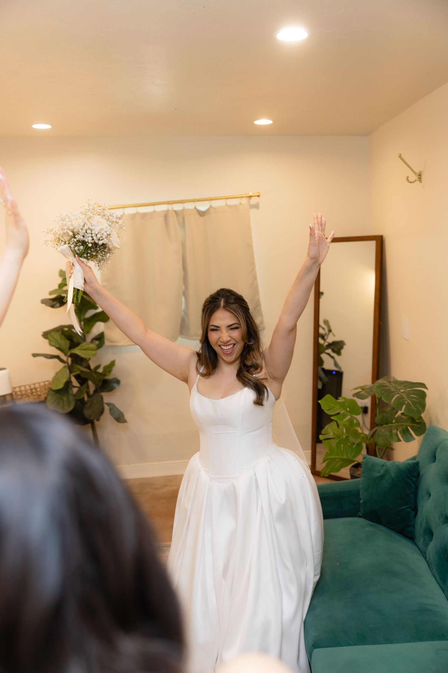 Excited bride in white wedding dress raises arms joyfully holding bouquet in bridal suite.