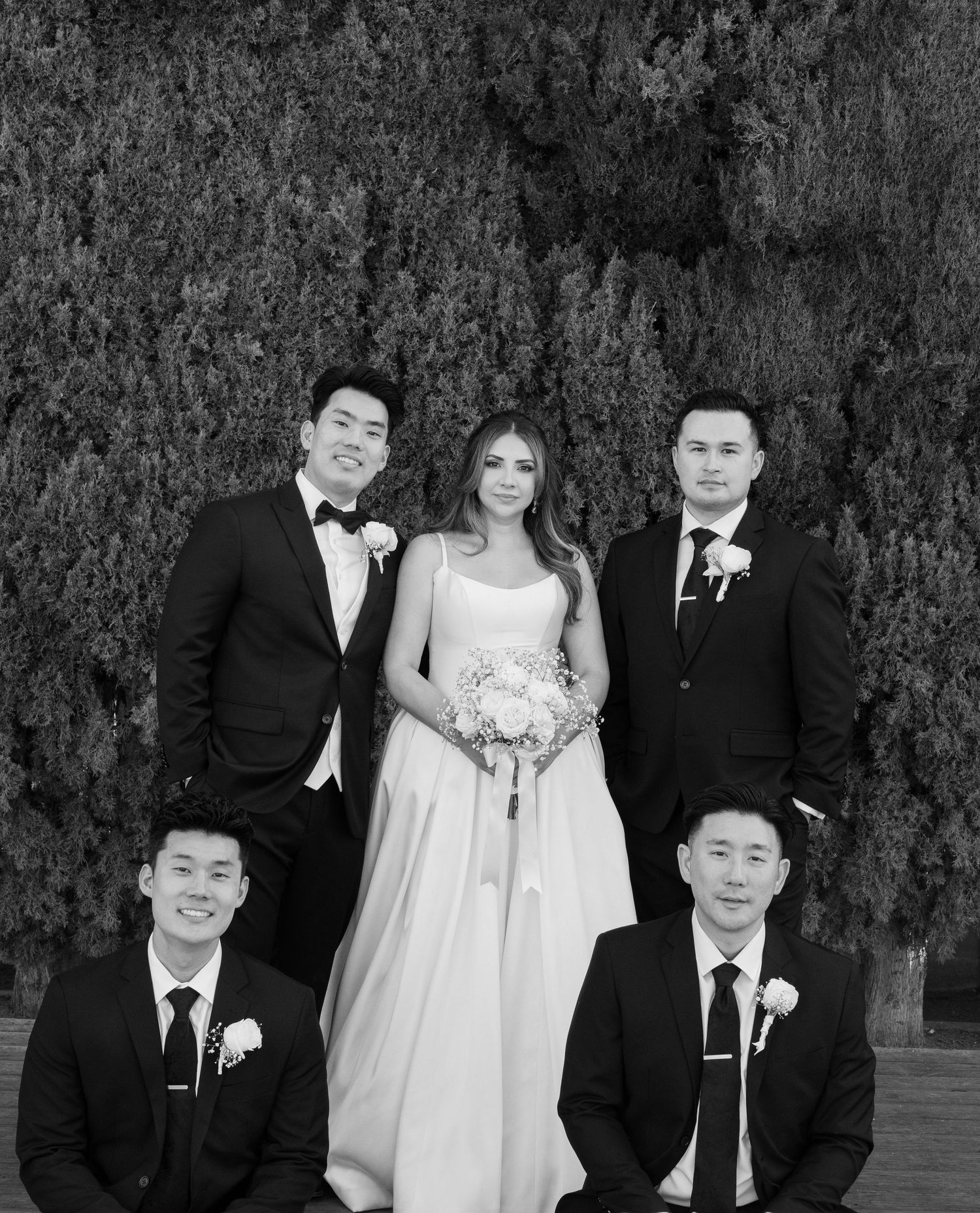 Black and white photo of bride with four groomsmen posing before a green hedge at wedding.