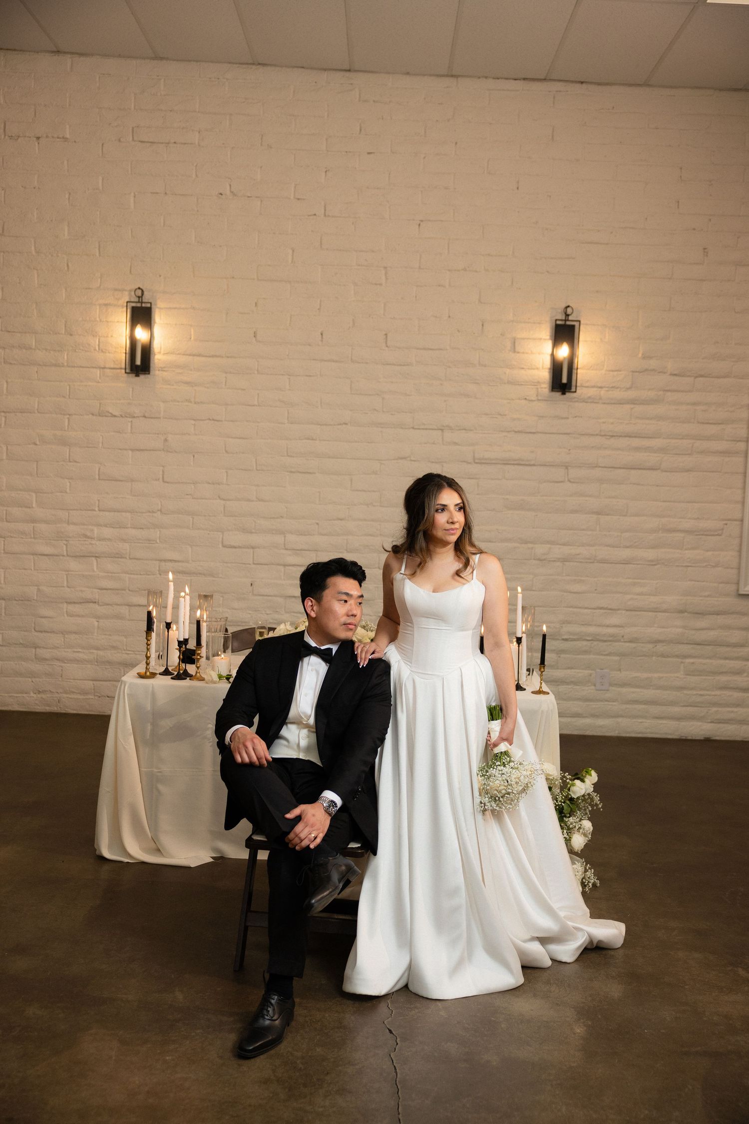Bride in white gown holding bouquet stands beside seated groom in black tuxedo before white brick wall.