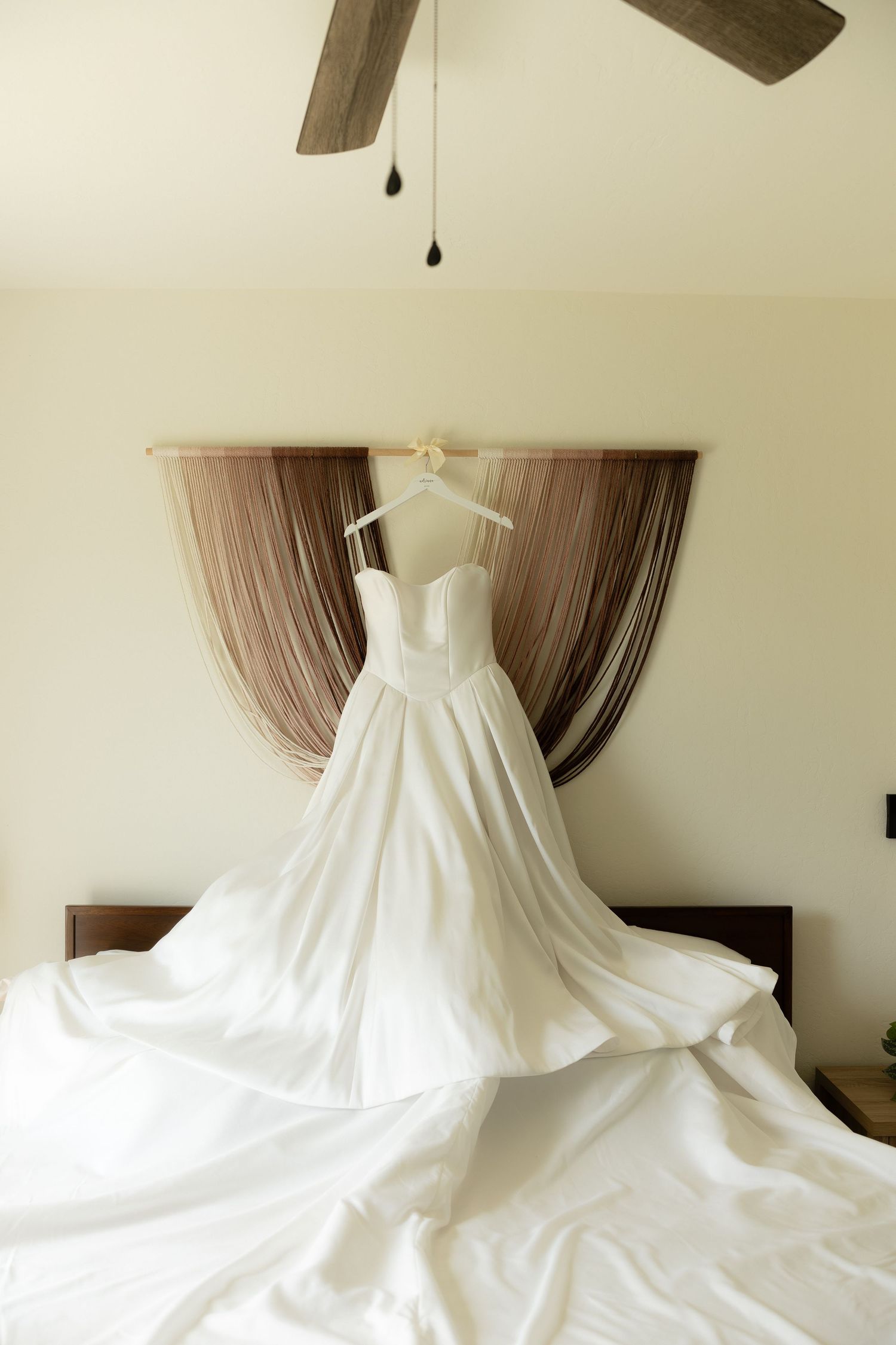 Elegant white ball gown wedding dress displayed on a hanger over a bed with flowing train in a bright bedroom.