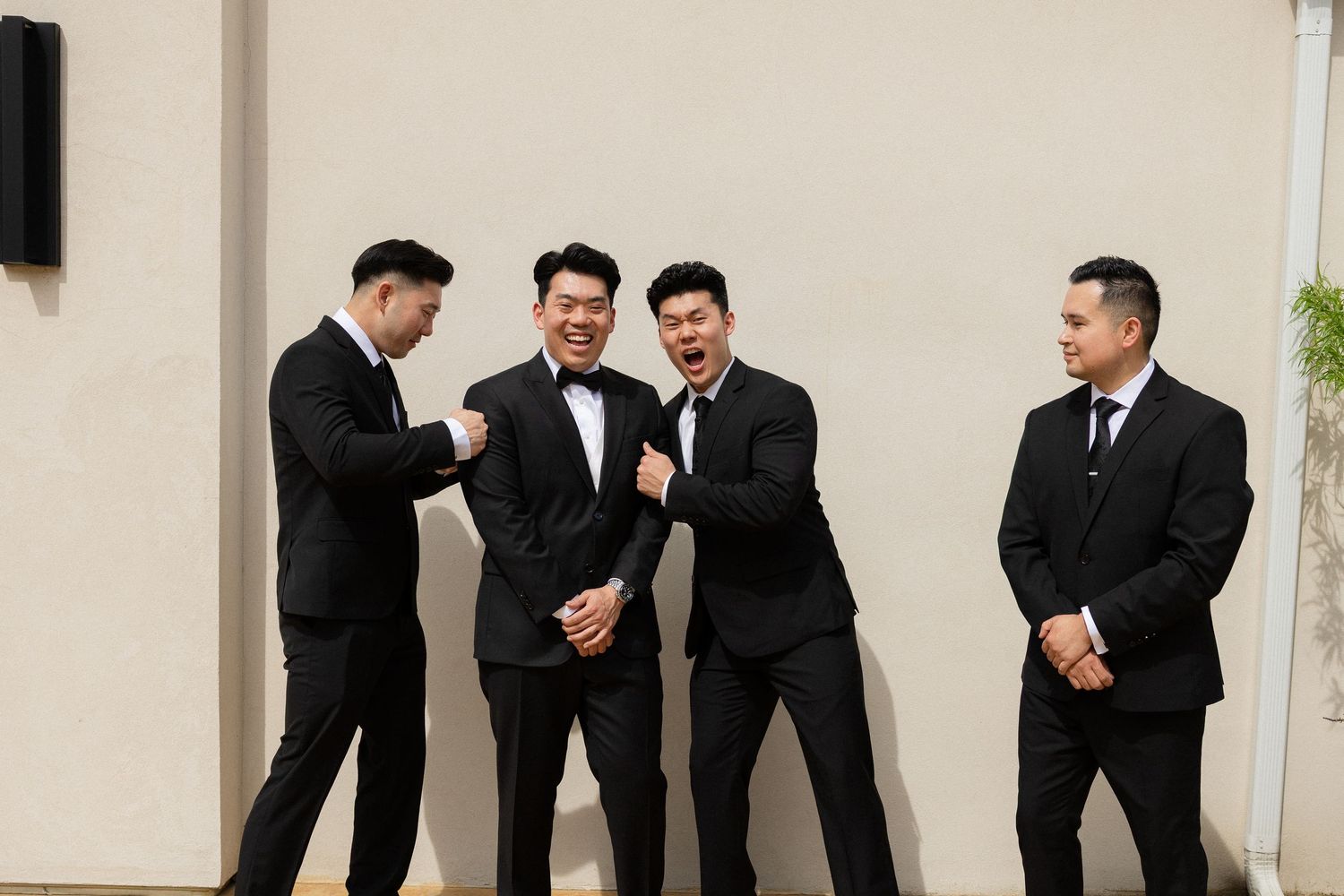 Four groomsmen in black suits laugh and pose together outdoors against a neutral wall before a wedding.