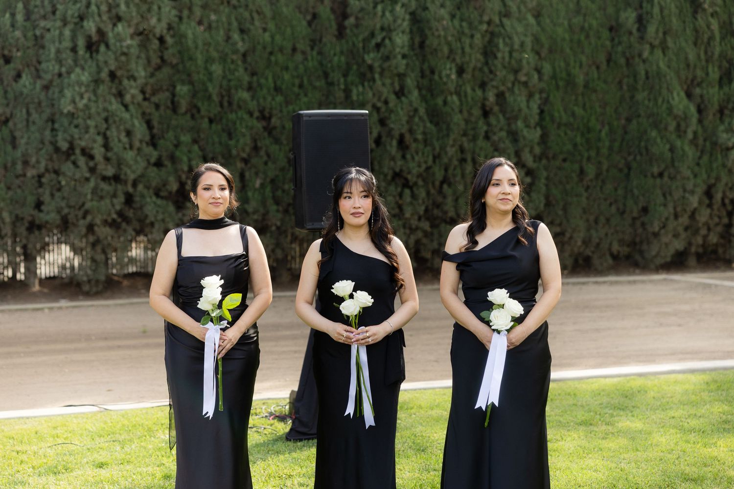 Three bridesmaids in black gowns holding white bouquets stand outdoors at a wedding ceremony.