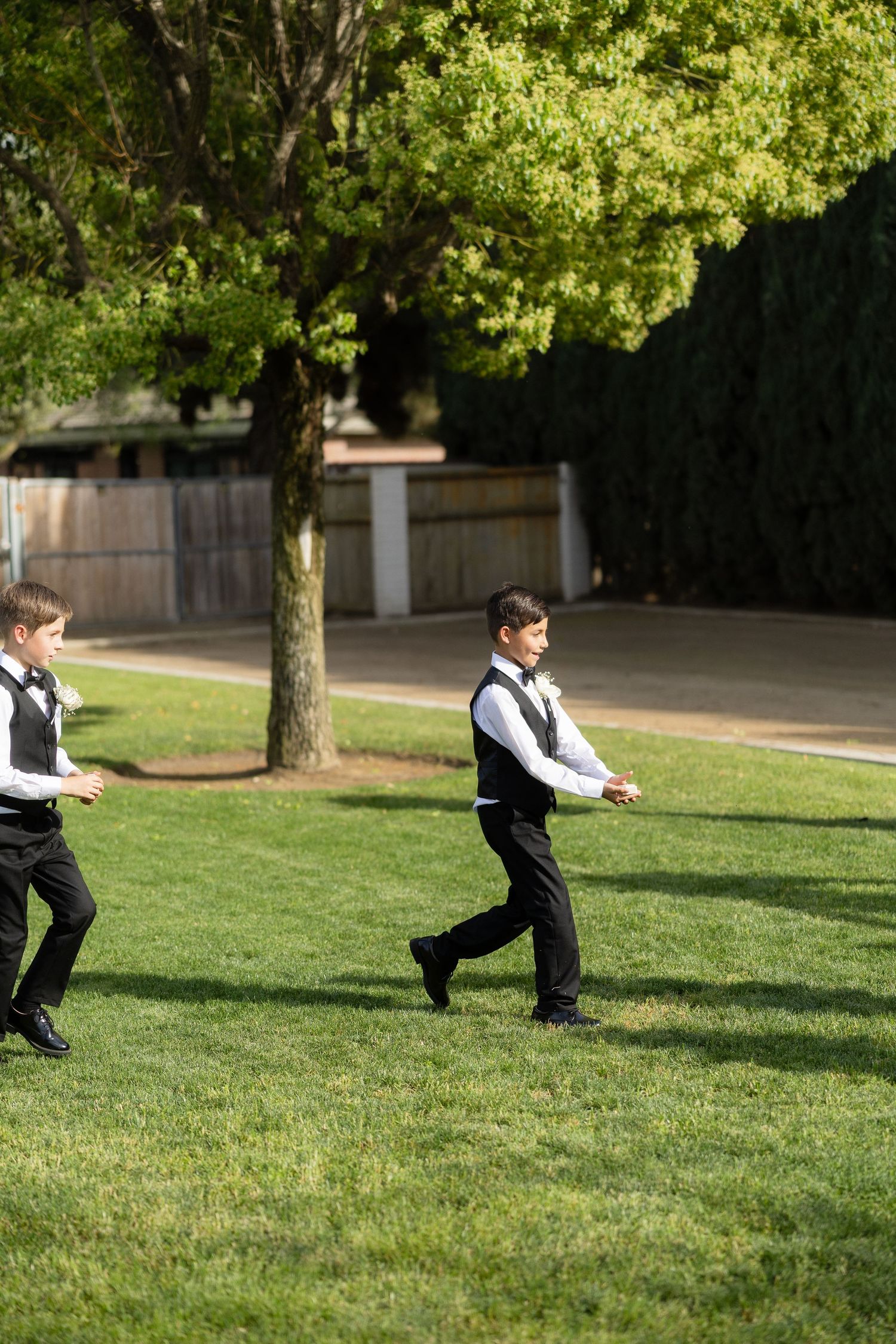 Two boys in black vests and pants walk across green lawn at outdoor wedding venue.