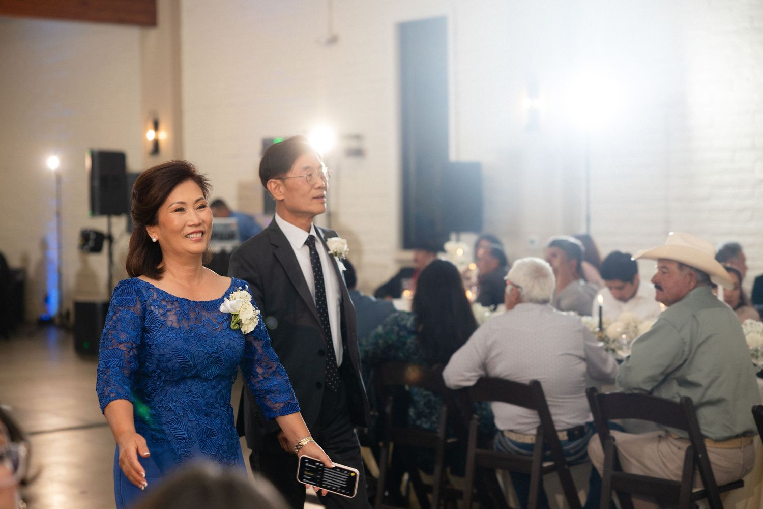 Elegantly dressed mother of groom in blue lace dress and father in black tuxedo walk into wedding reception hall together.