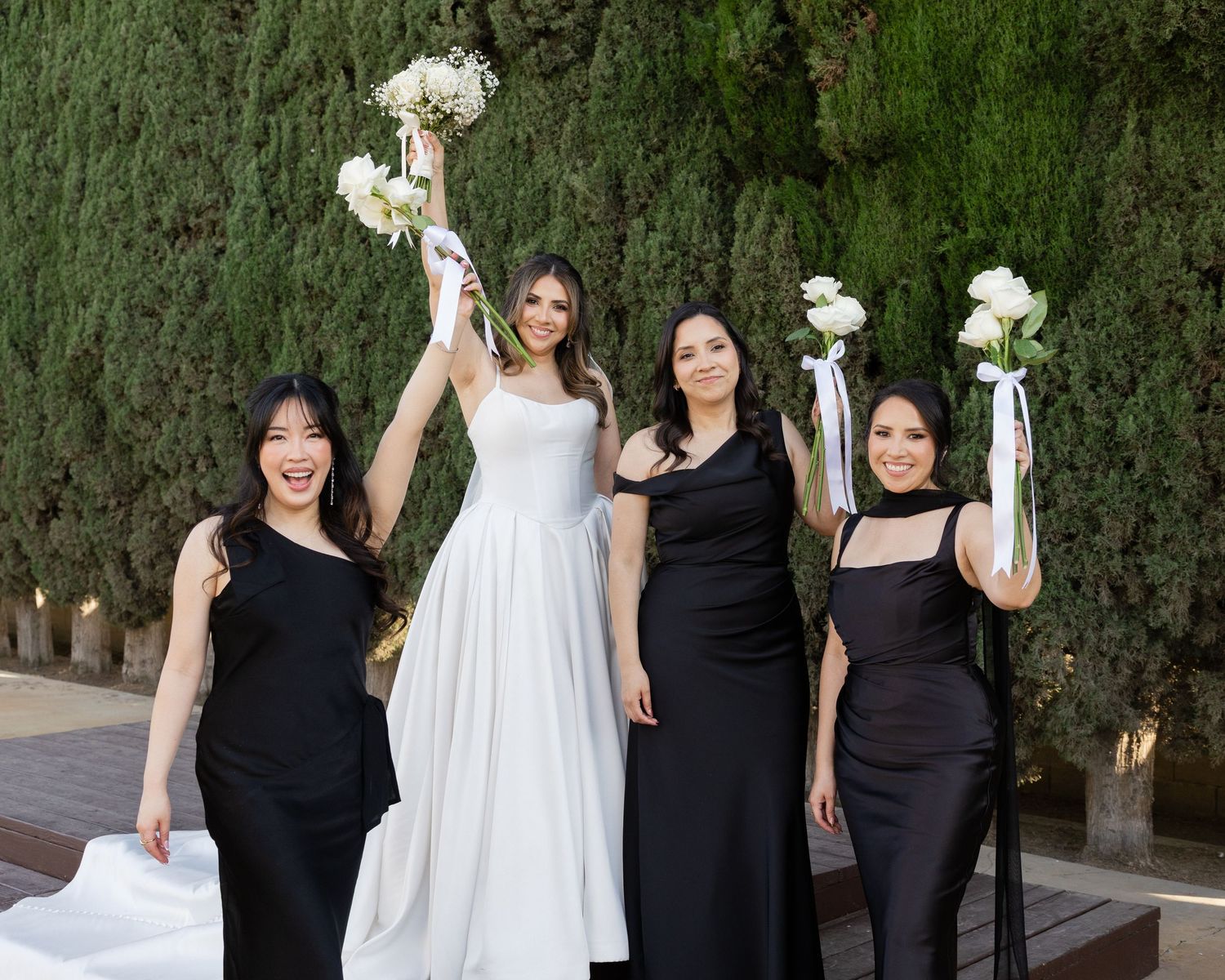 Bride in white gown with three bridesmaids in black dresses holding white bouquets aloft outdoors.