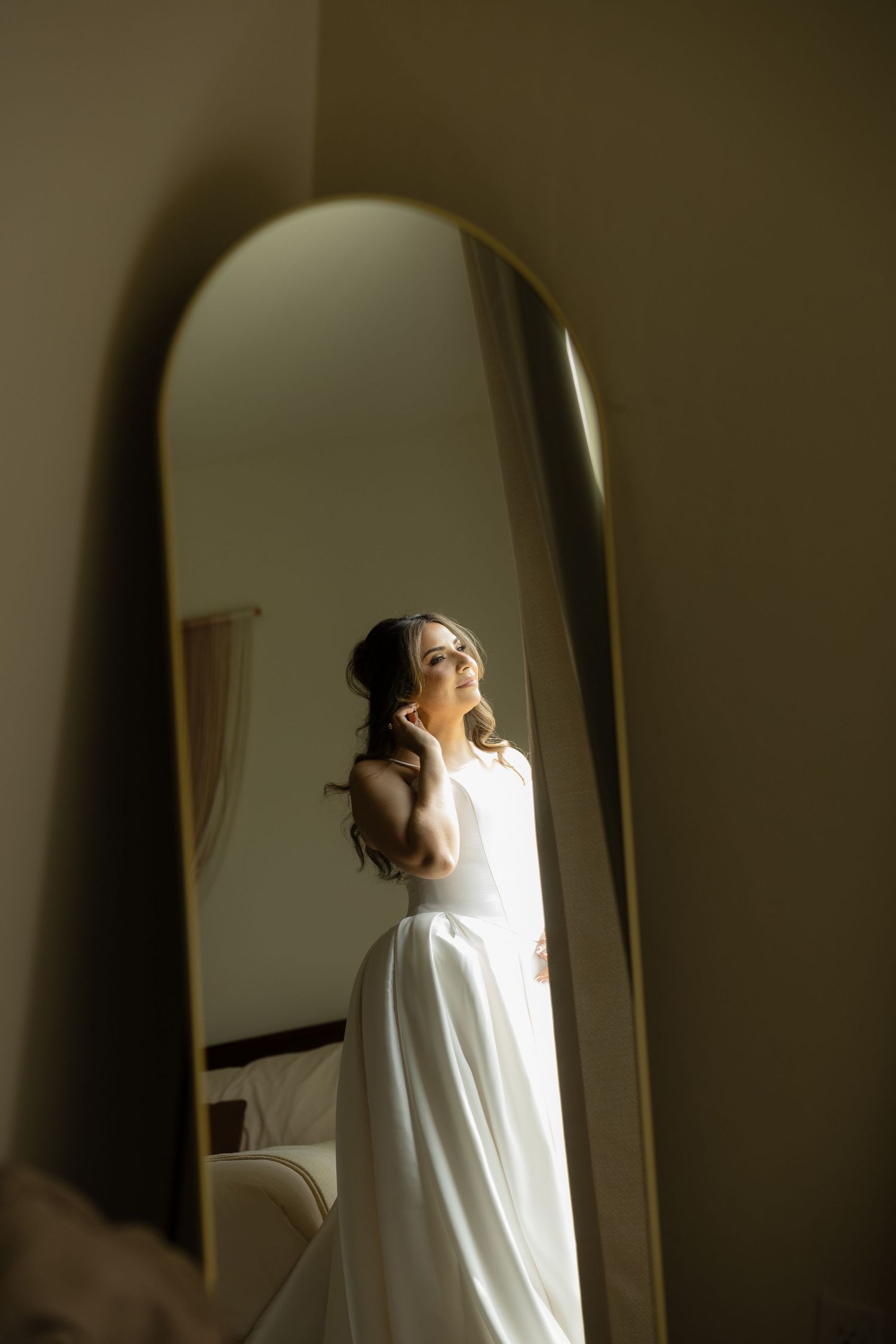 Elegant bride in a white gown admiring herself in a large arched mirror before her wedding.