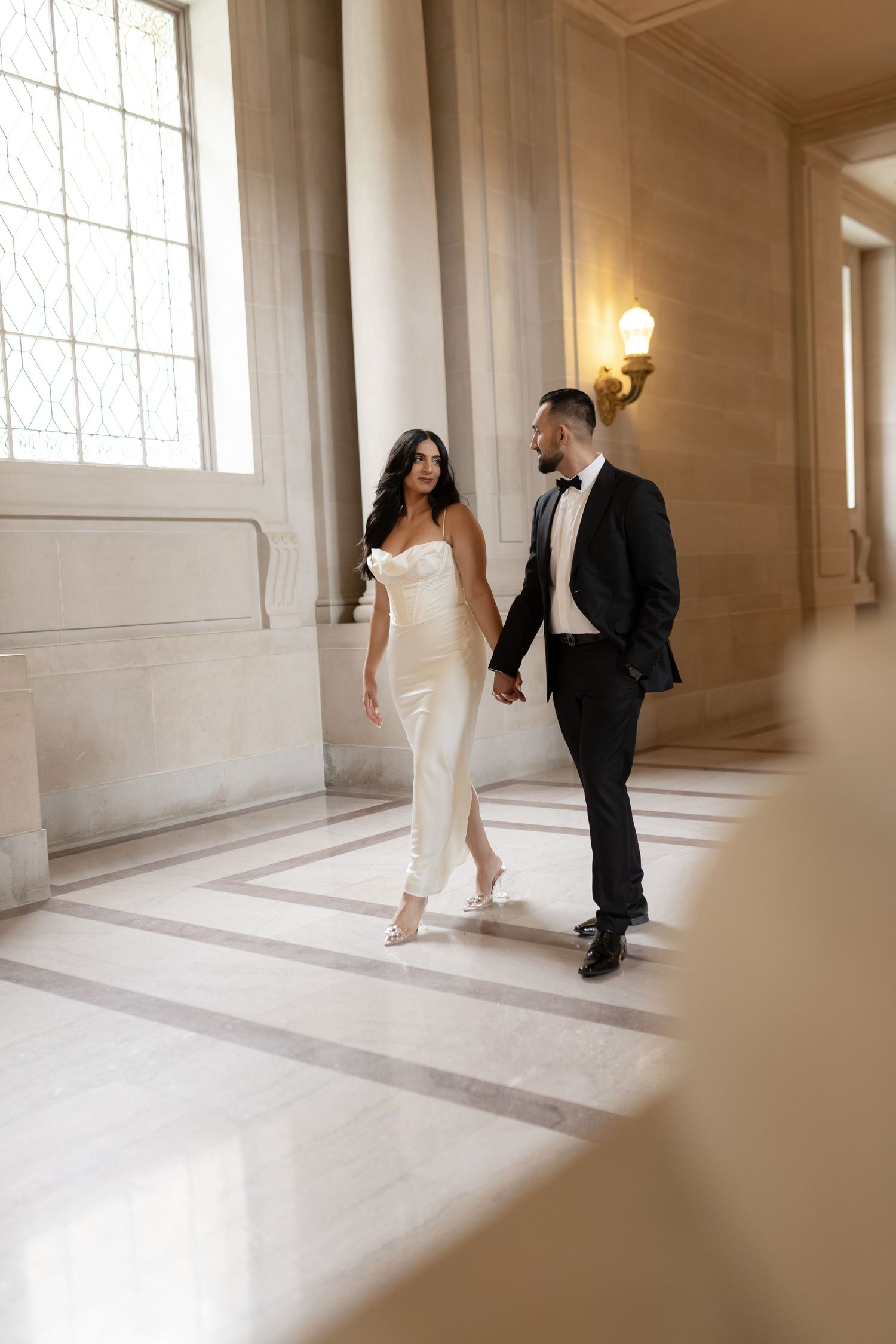 A couple in formal wedding attire shares an intimate moment in a bright, minimalist hallway with natural light streaming in.