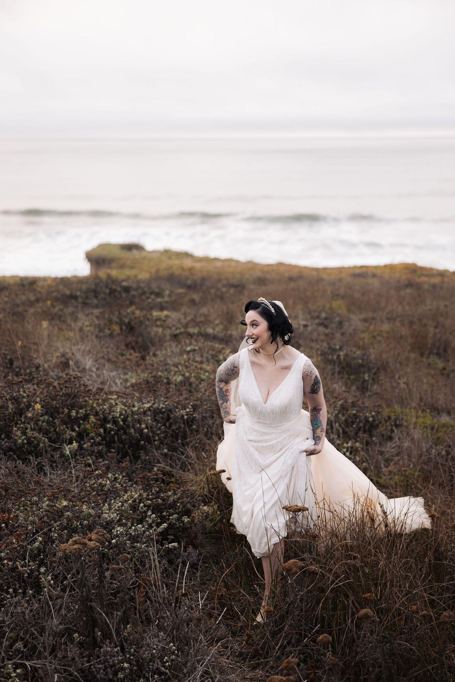 A figure in a flowing white dress sits gracefully among wild coastal grasses on a moody, overcast day.