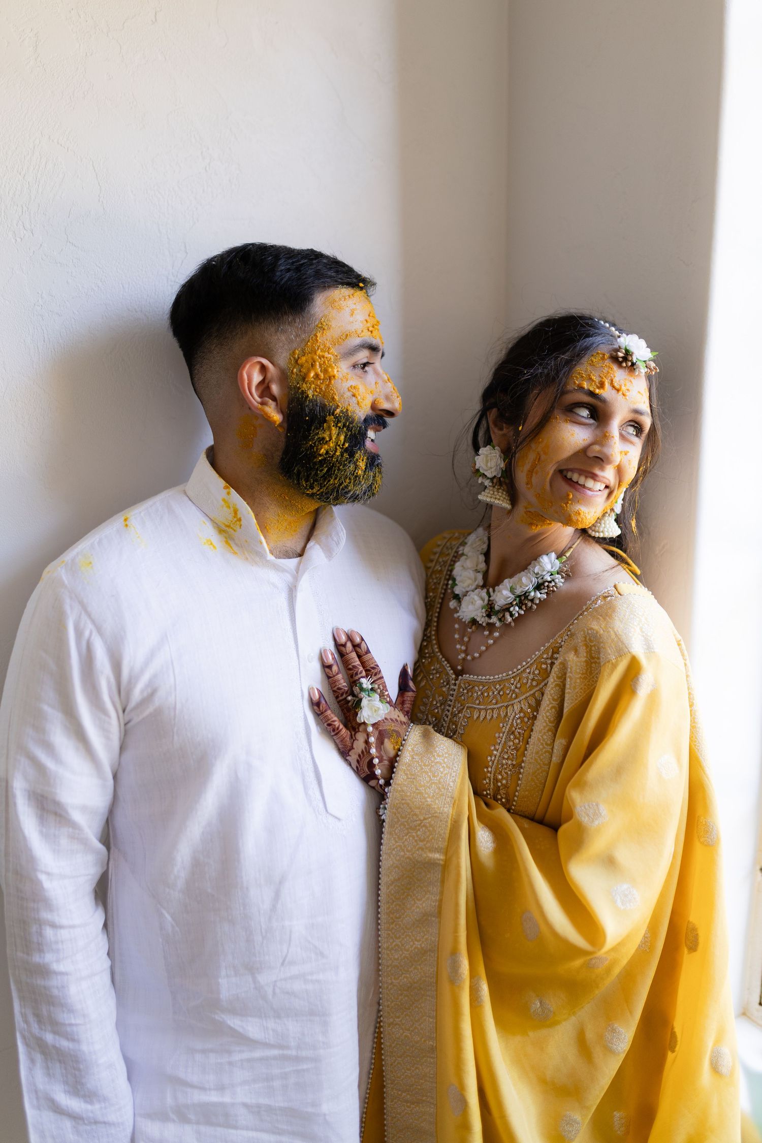 Couple in yellow and white traditional dress pose together against white wall in romantic photoshoot.