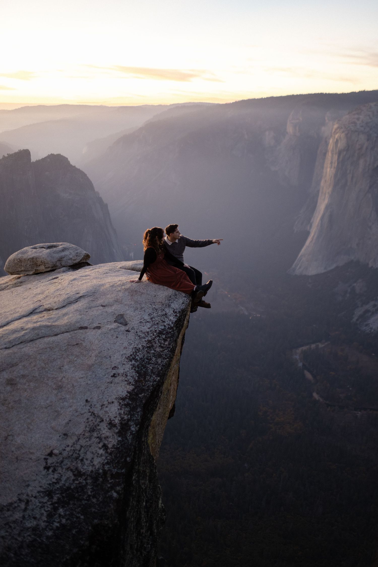 Two people sitting precariously on edge of cliff overlooking vast mountain valley.