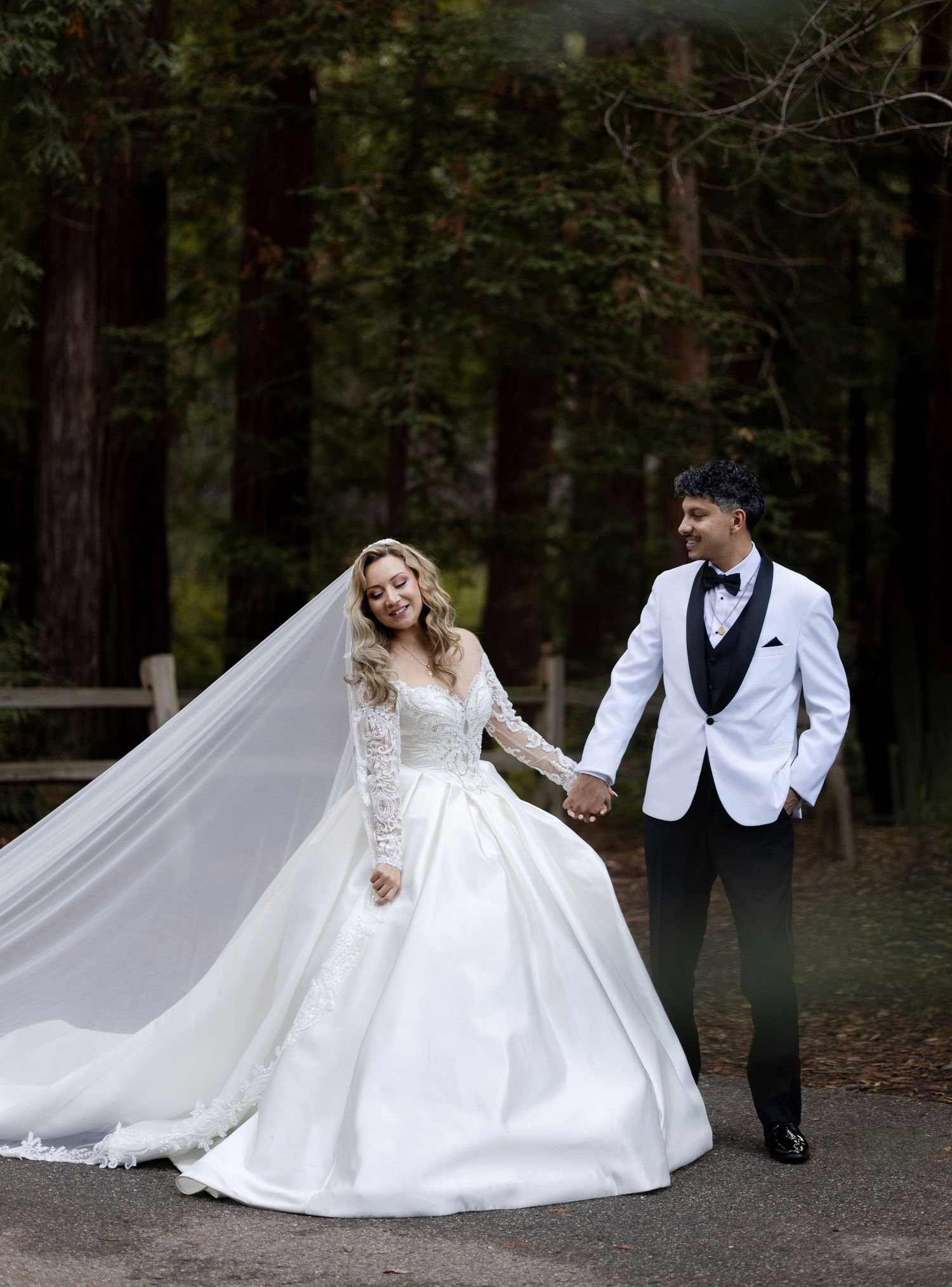 A joyful wedding couple poses on a forest path, with the bride's veil flowing in the breeze.
