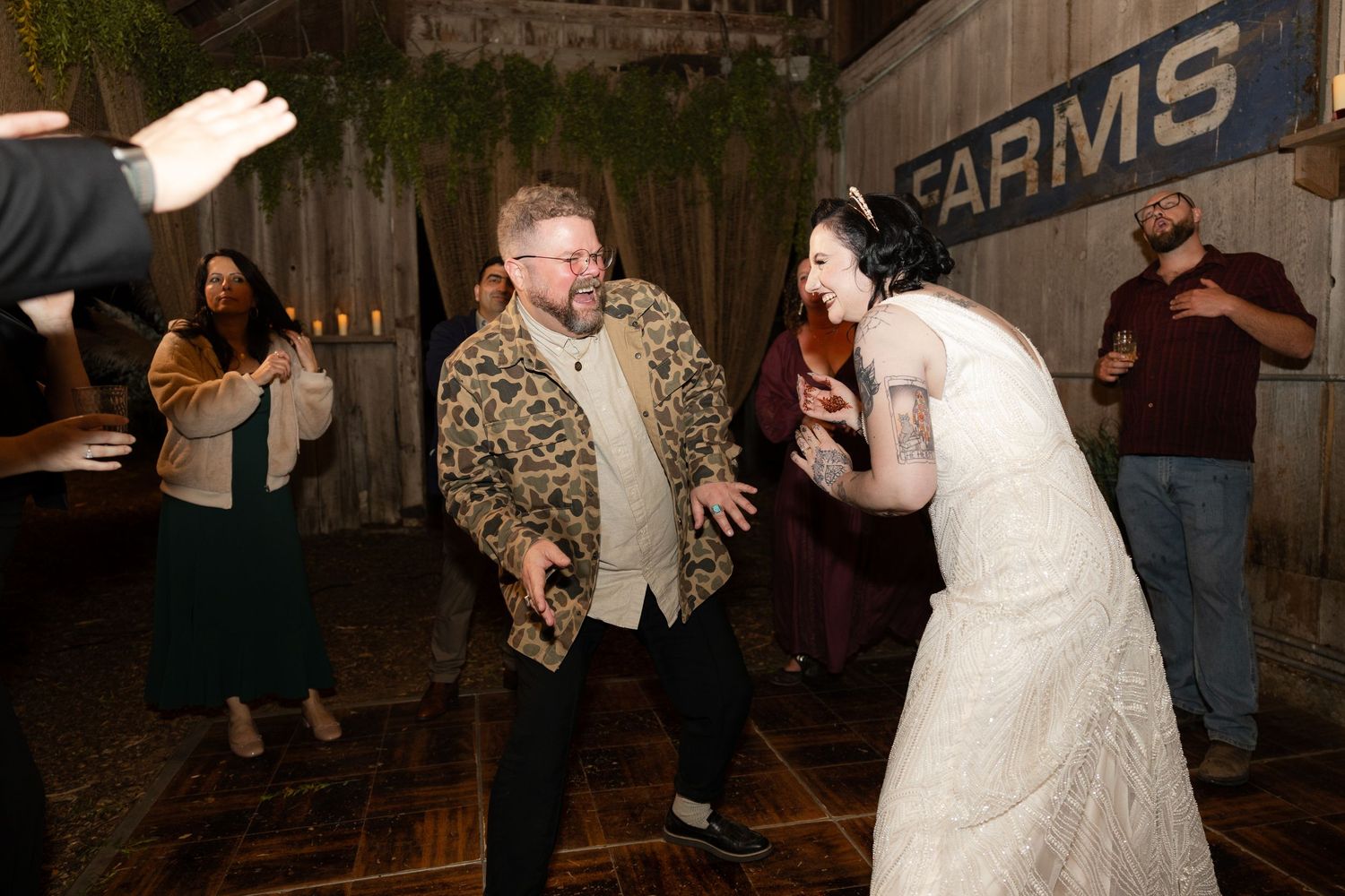 Guests dancing at a rustic barn wedding reception with 'ARMS' signage visible on the wall.