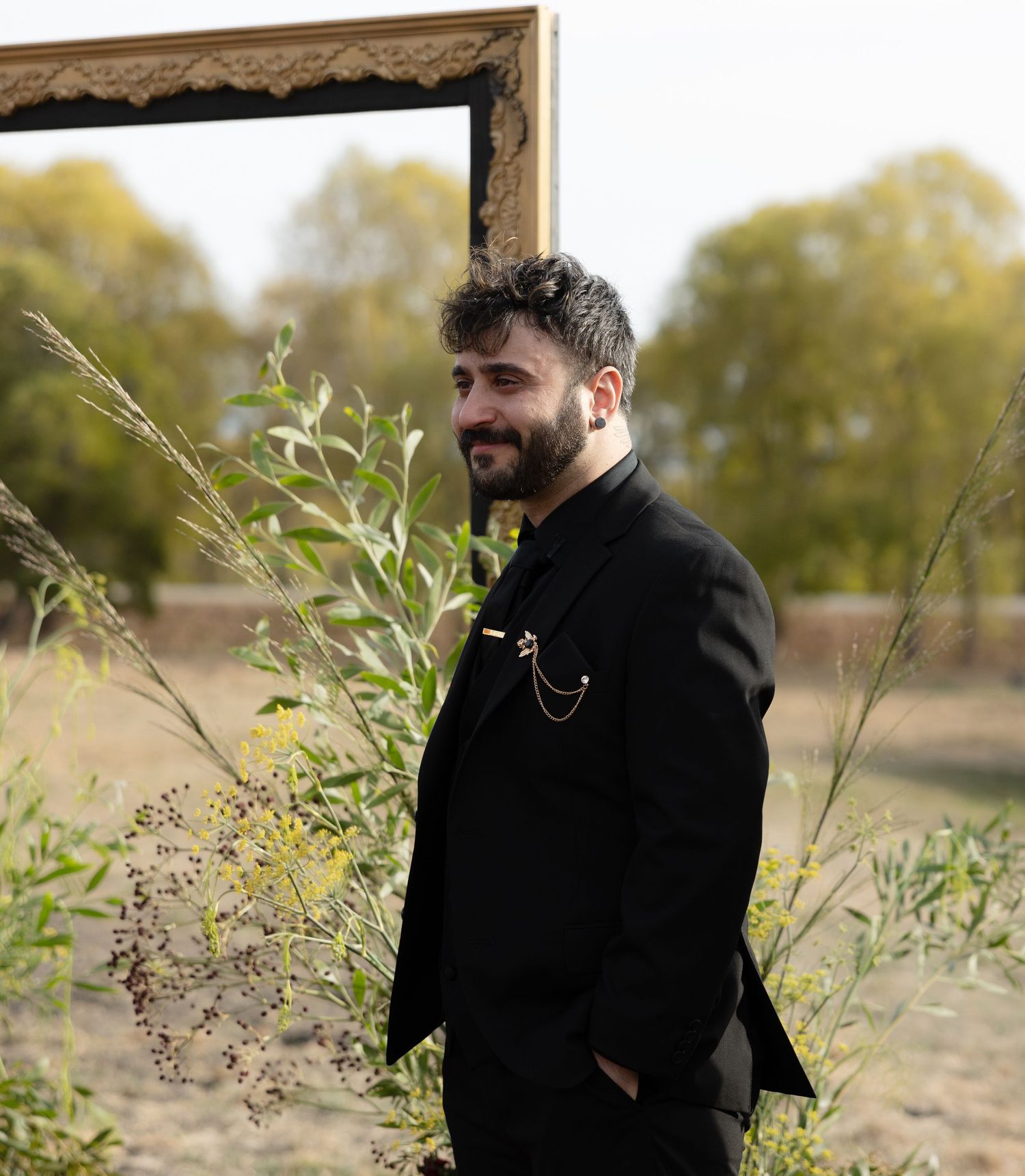 Person in black formal attire stands thoughtfully in an autumn field setting.