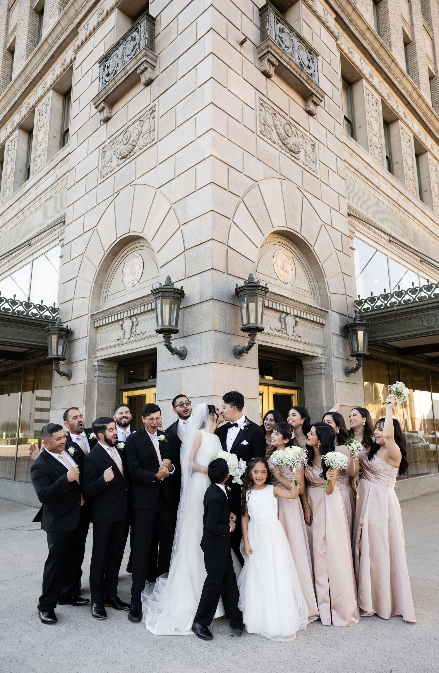 Large wedding party gathered for formal photos against stone building backdrop.