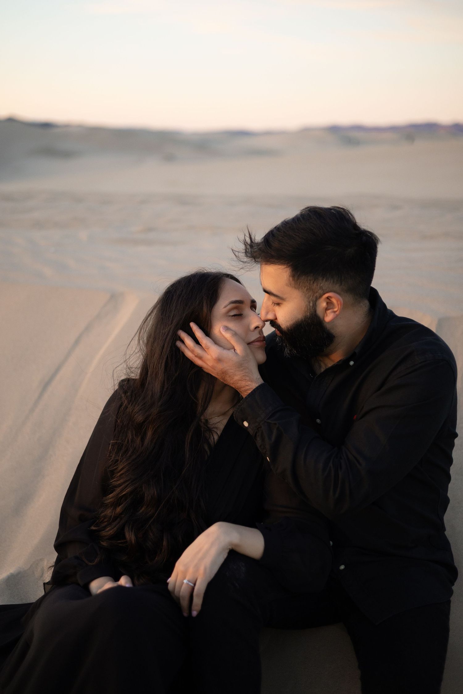A romantic couple in black attire shares intimate moments against a desert landscape during golden hour.